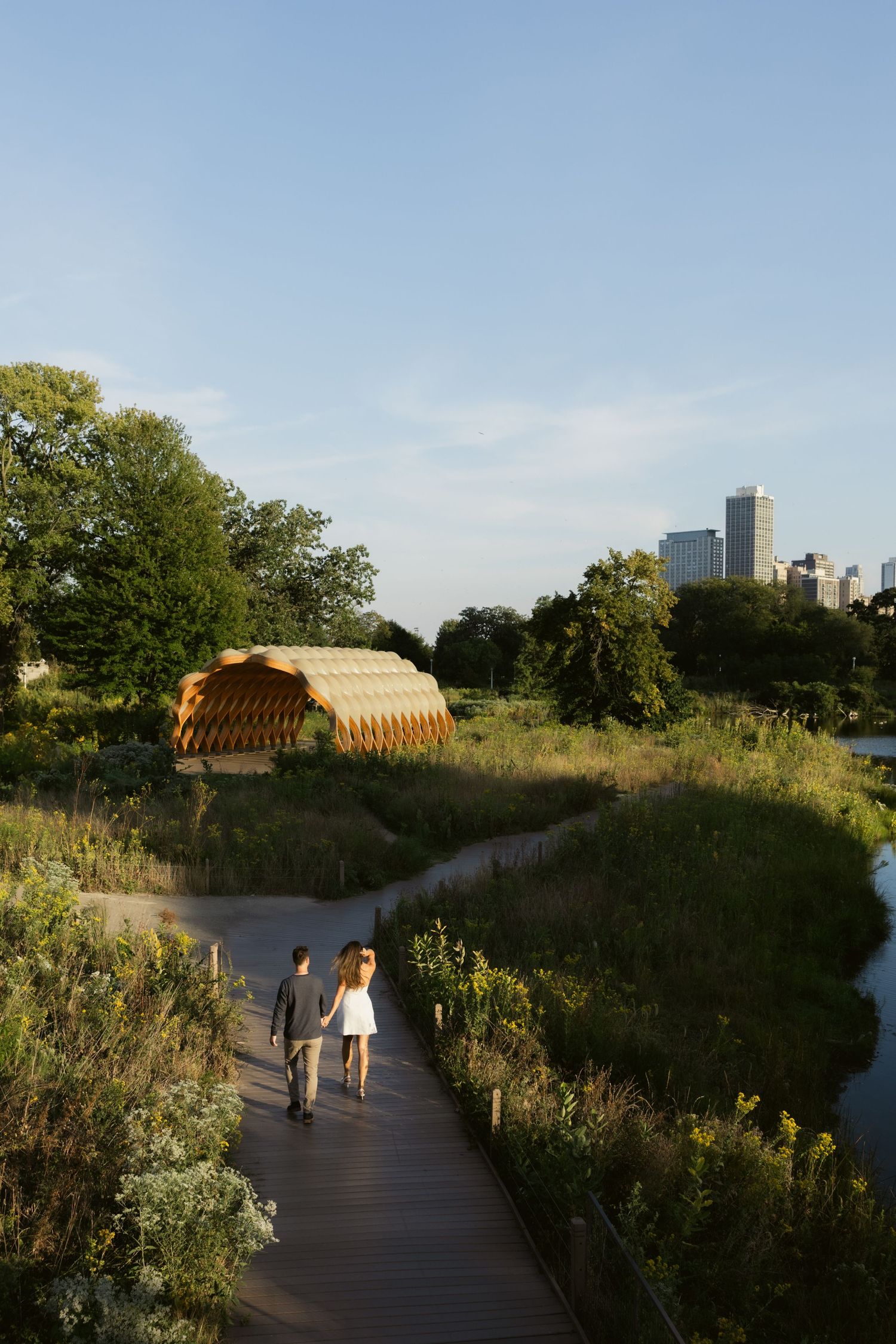 Two people walking on wooden path through natural area with city views.