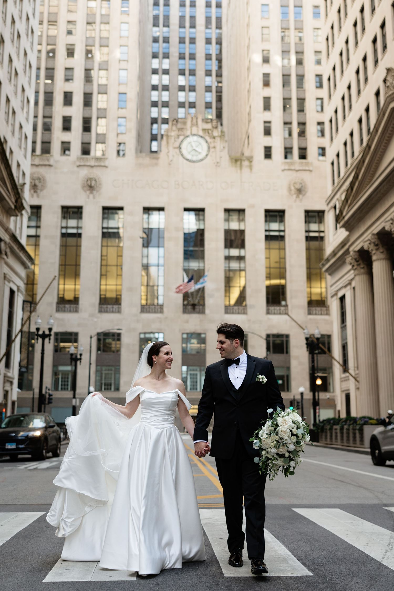 A wedding couple walks hand in hand on a city street with an elegant art deco building and clock tower in the background.
