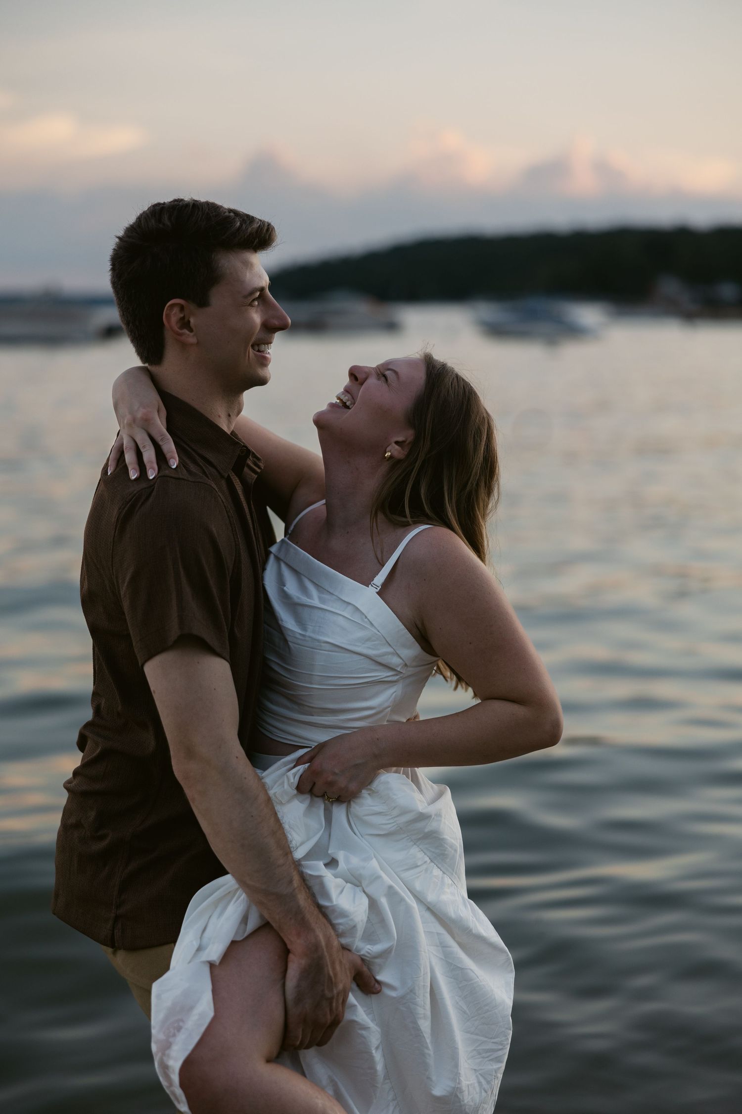 A romantic couple embraces in the water during sunset with boats in the distance.