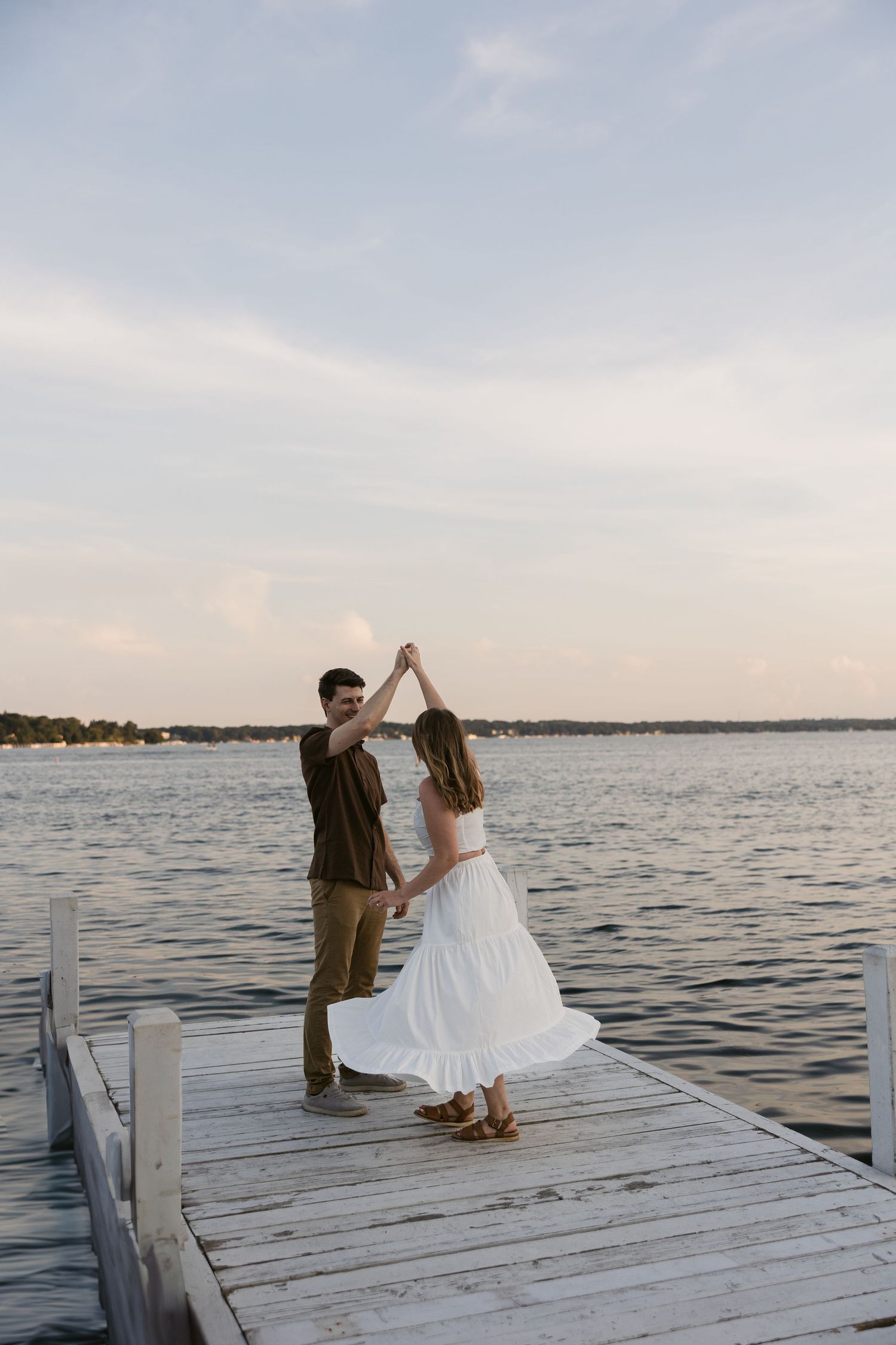 A couple sits together on a wooden dock by the water during sunset.