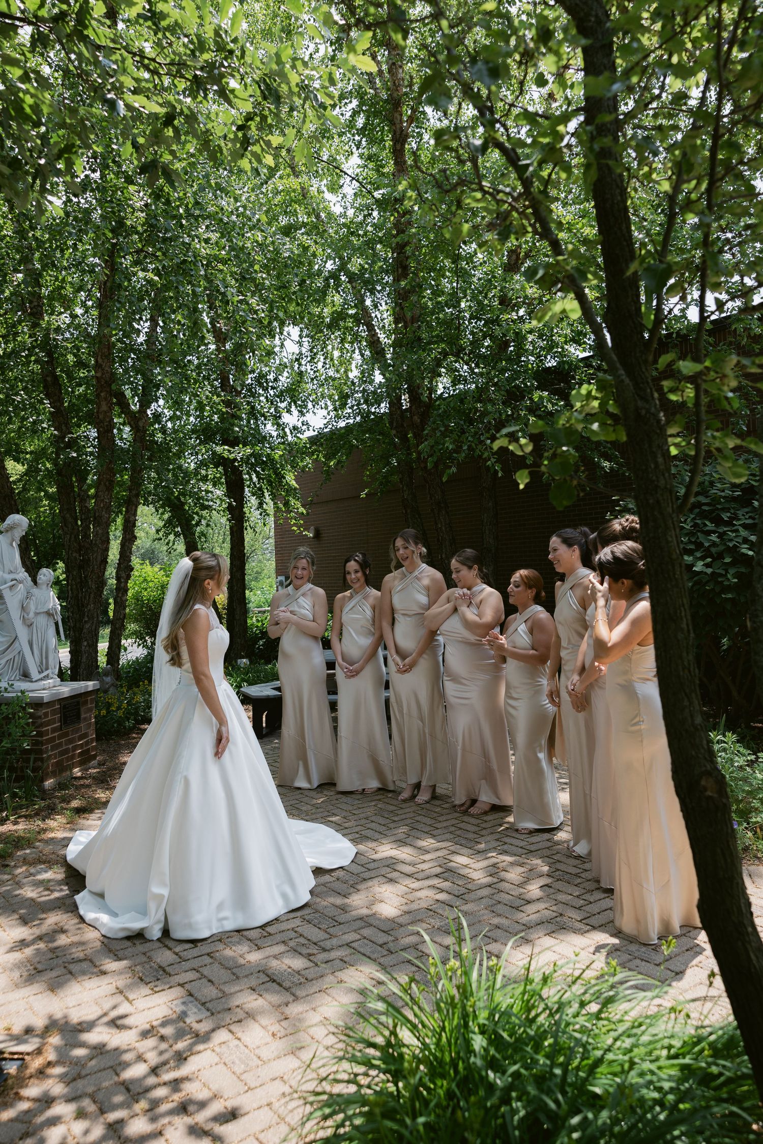 A bride in a white gown stands with bridesmaids wearing beige dresses on a garden path surrounded by trees.