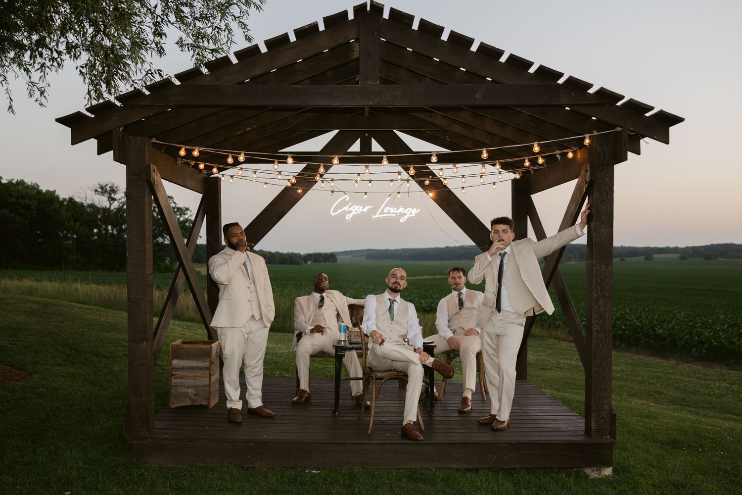 Group in white attire sits on rustic porch swing during outdoor wedding celebration at sunset.