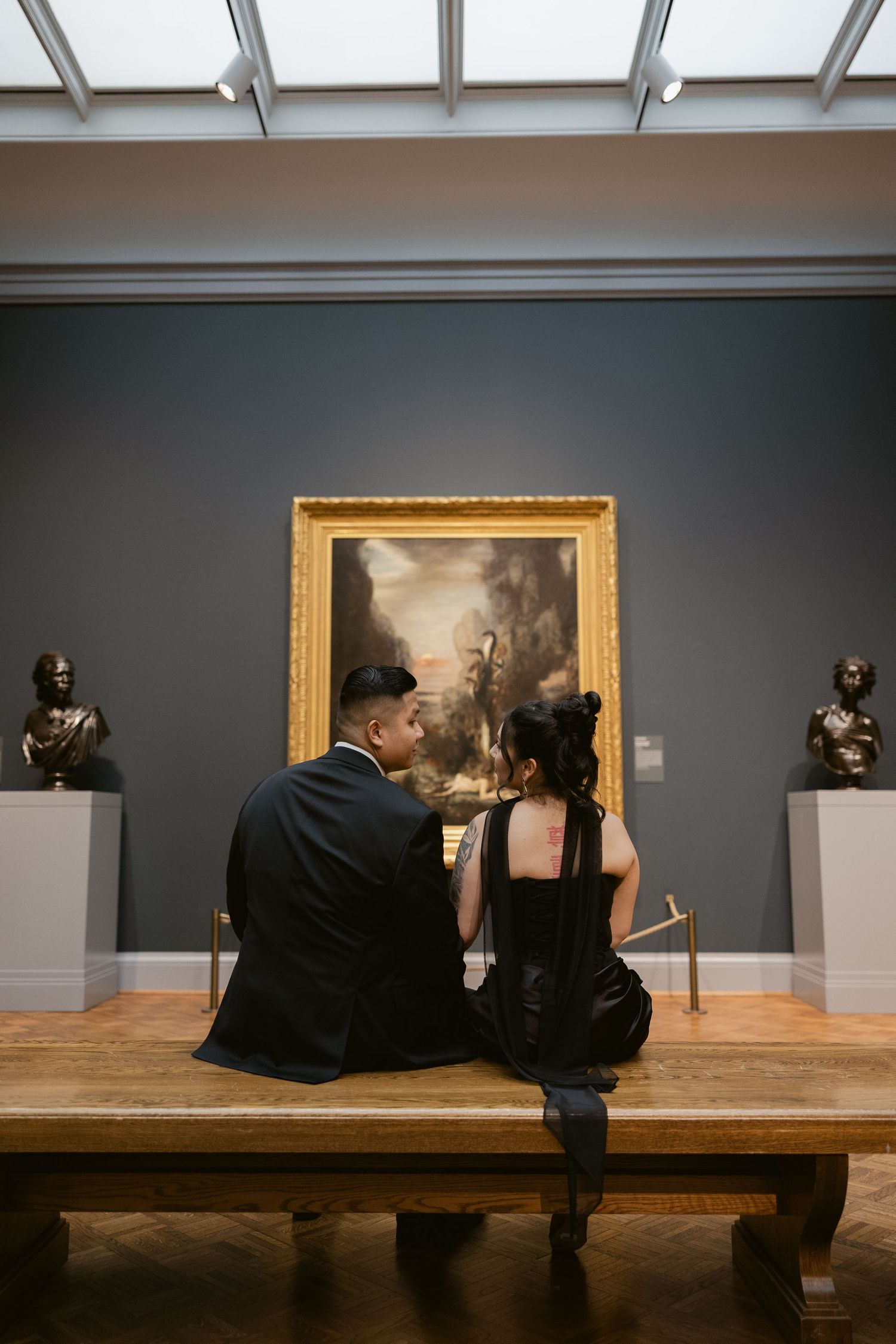 A couple sits on a wooden bench viewing artwork in an elegant museum gallery with dark walls.