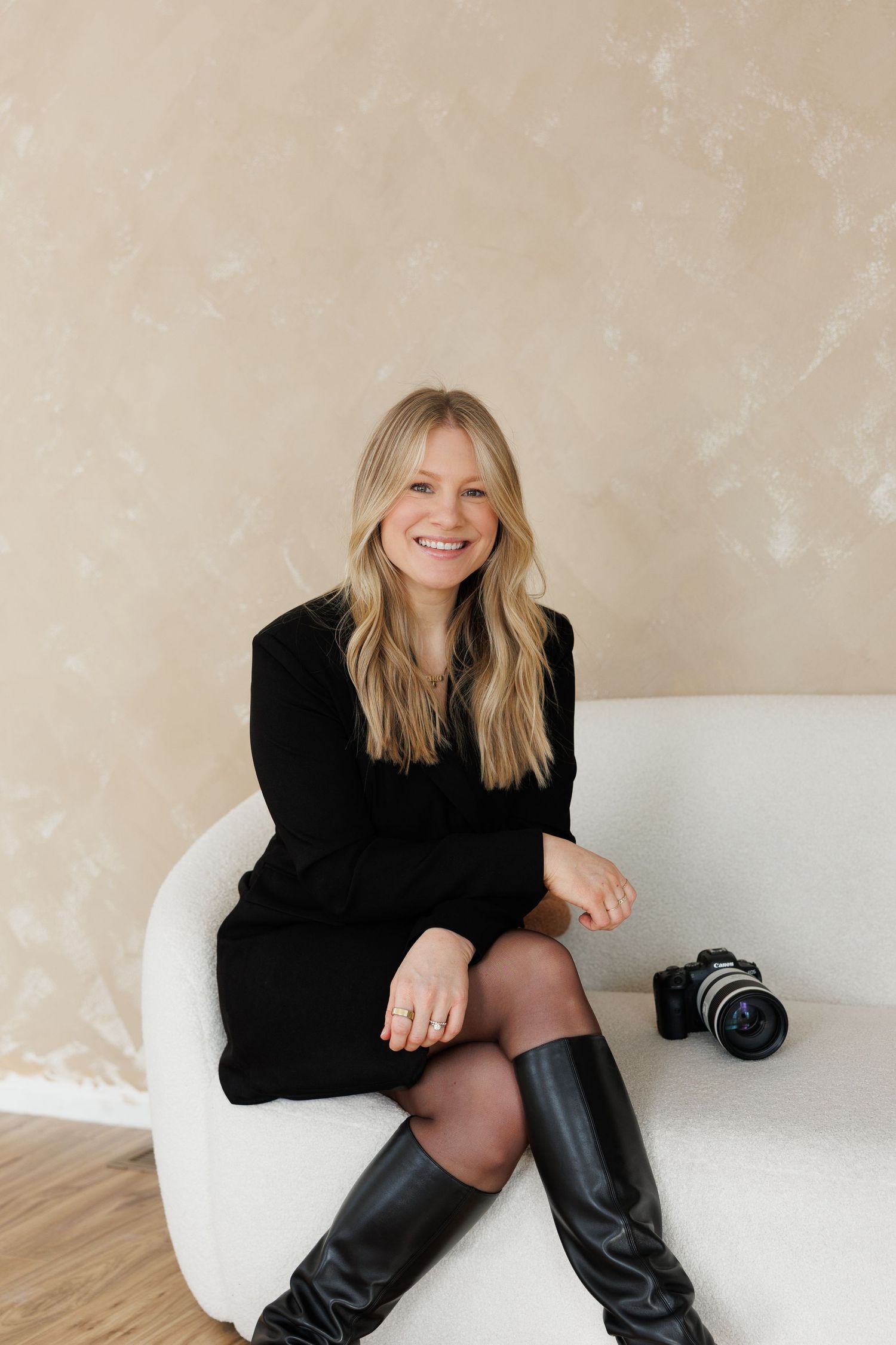 A person in a black sweater and boots poses casually on a white surface against a beige textured wall.