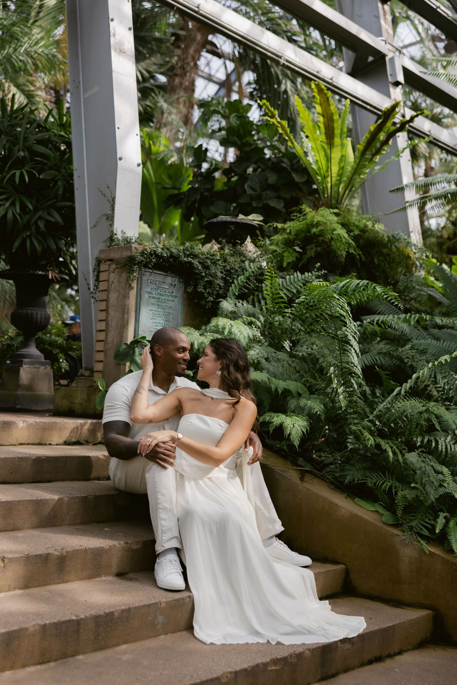 A couple in white attire shares an intimate moment on stone steps surrounded by lush tropical greenery and plants.