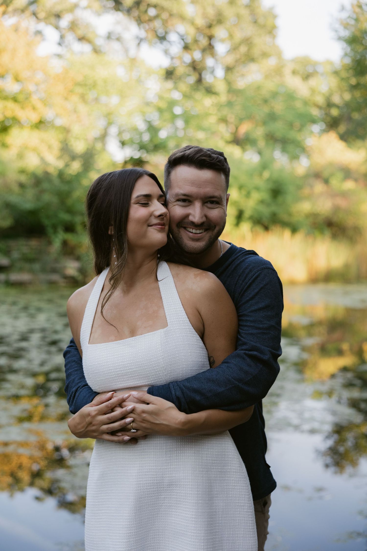 Couple embraces lovingly beside a tranquil pond with water lilies and trees in the background.