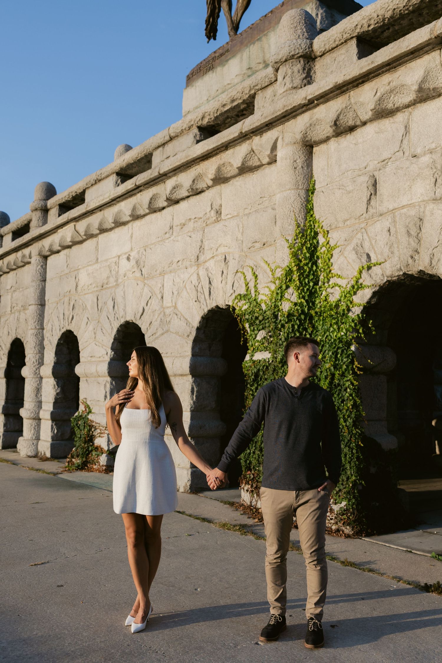 A romantic couple strolling along an old stone wall with arched windows.