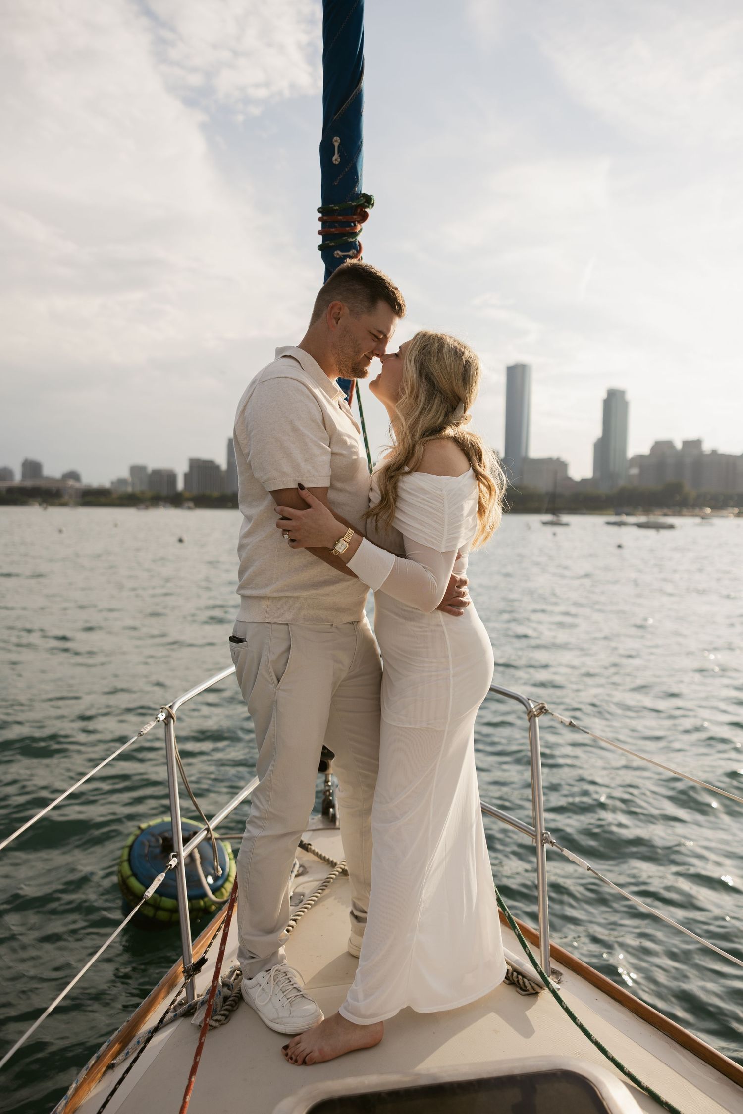 A couple in matching white outfits share an intimate moment on the bow of a sailboat with a city skyline backdrop.