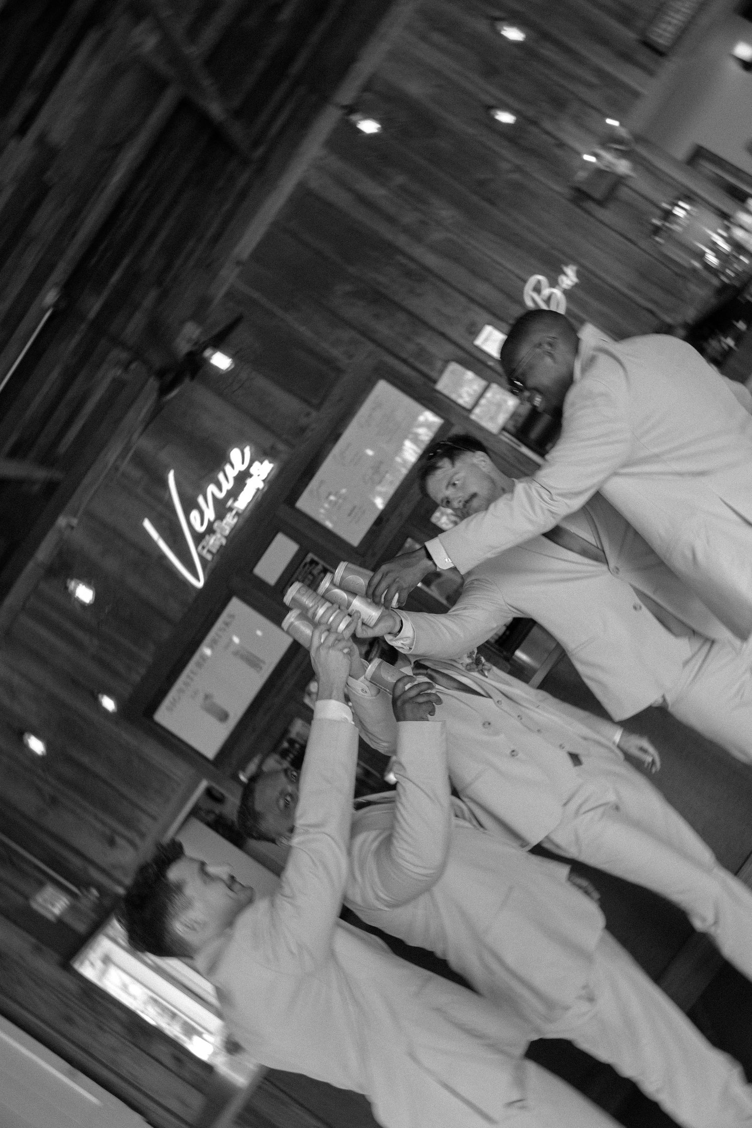 Bartender in light colored attire pours drinks at a venue with illuminated decor in black and white photography.