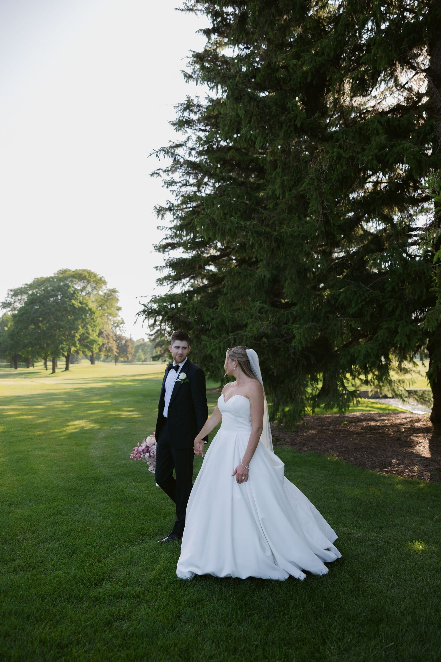 A wedding couple poses together on a lush green lawn with tall evergreen trees in the background.