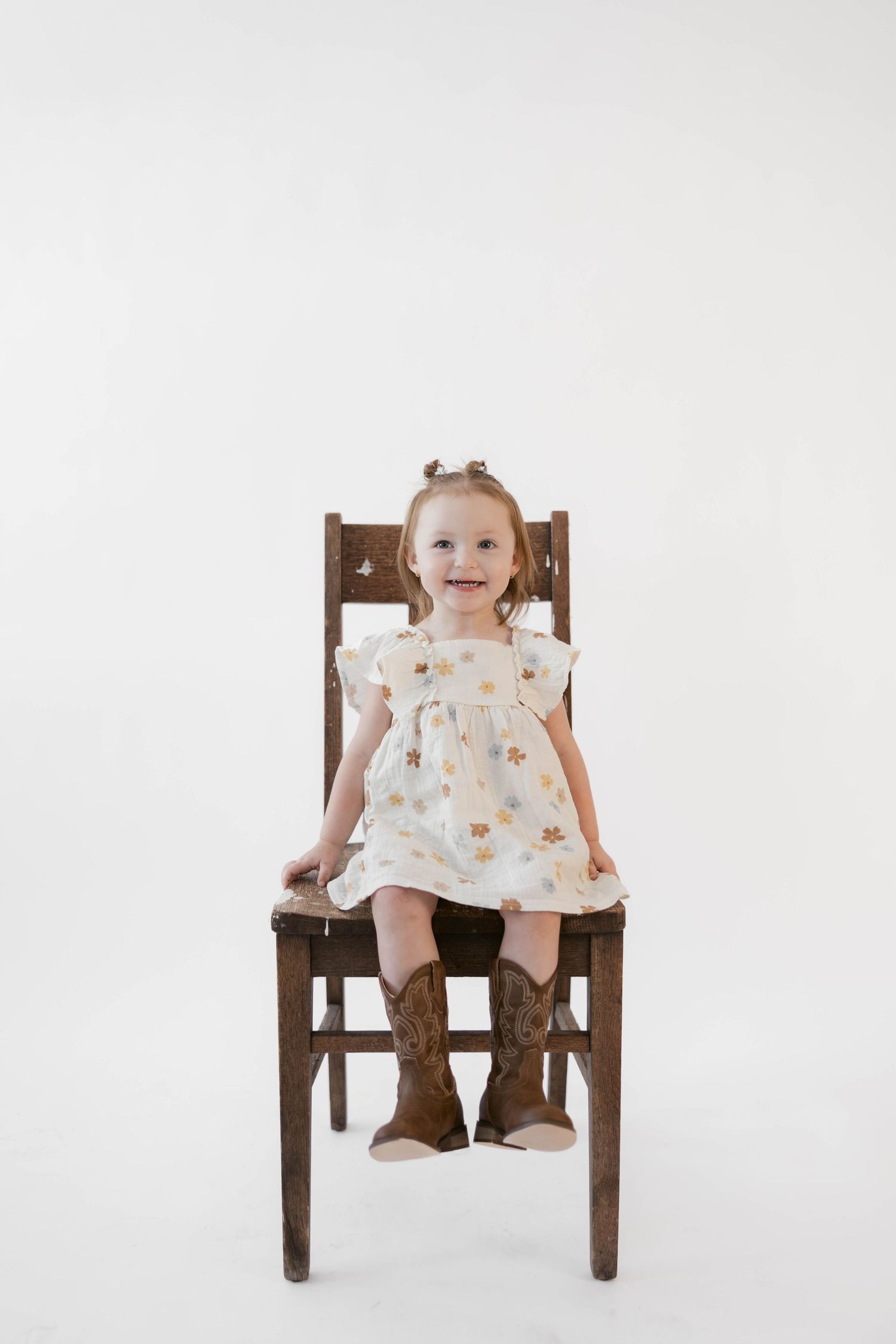 Smiling toddler in a floral dress and cowboy boots sitting on a wooden chair.