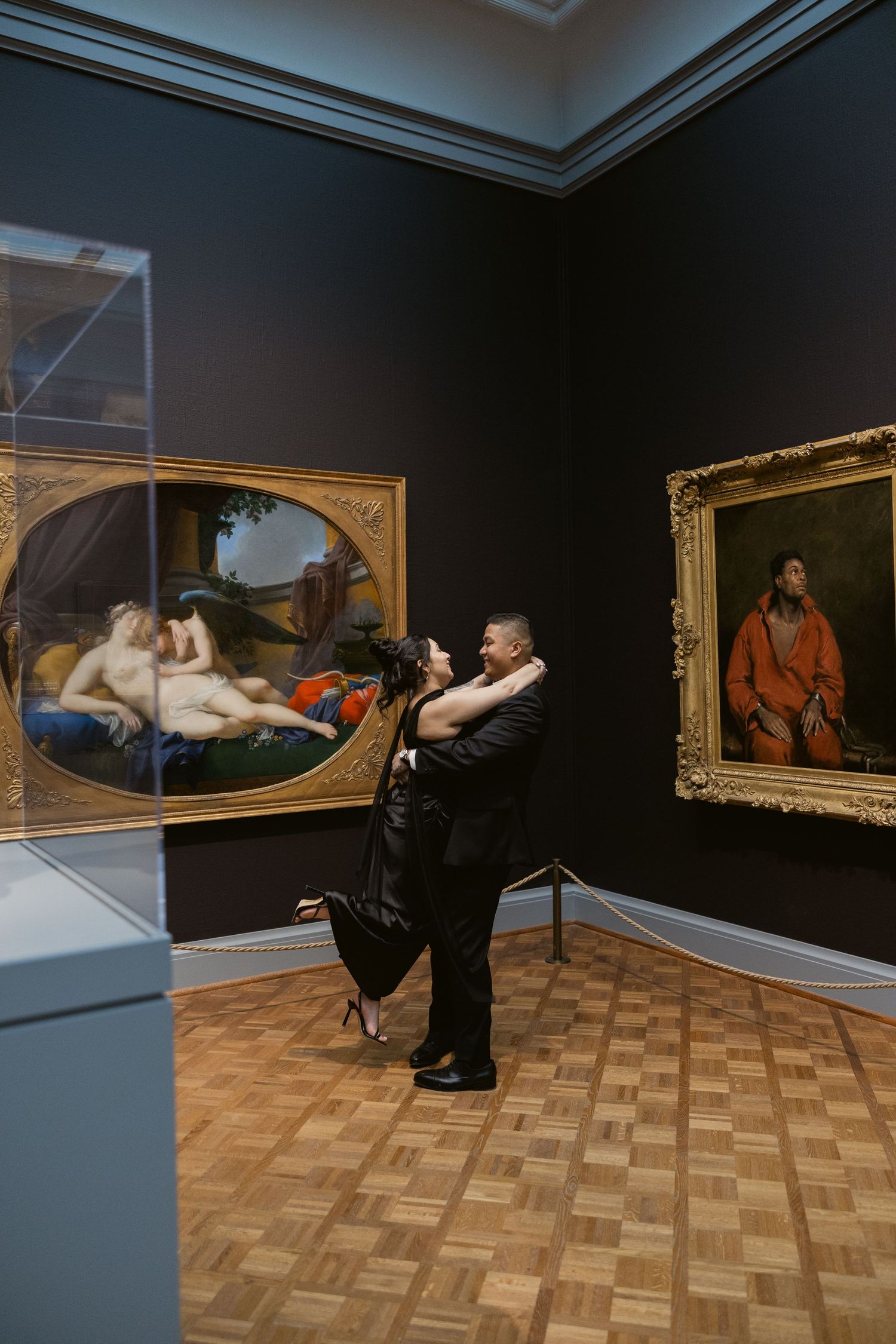 Two people in formal wear pose artistically in an art gallery with classical paintings.