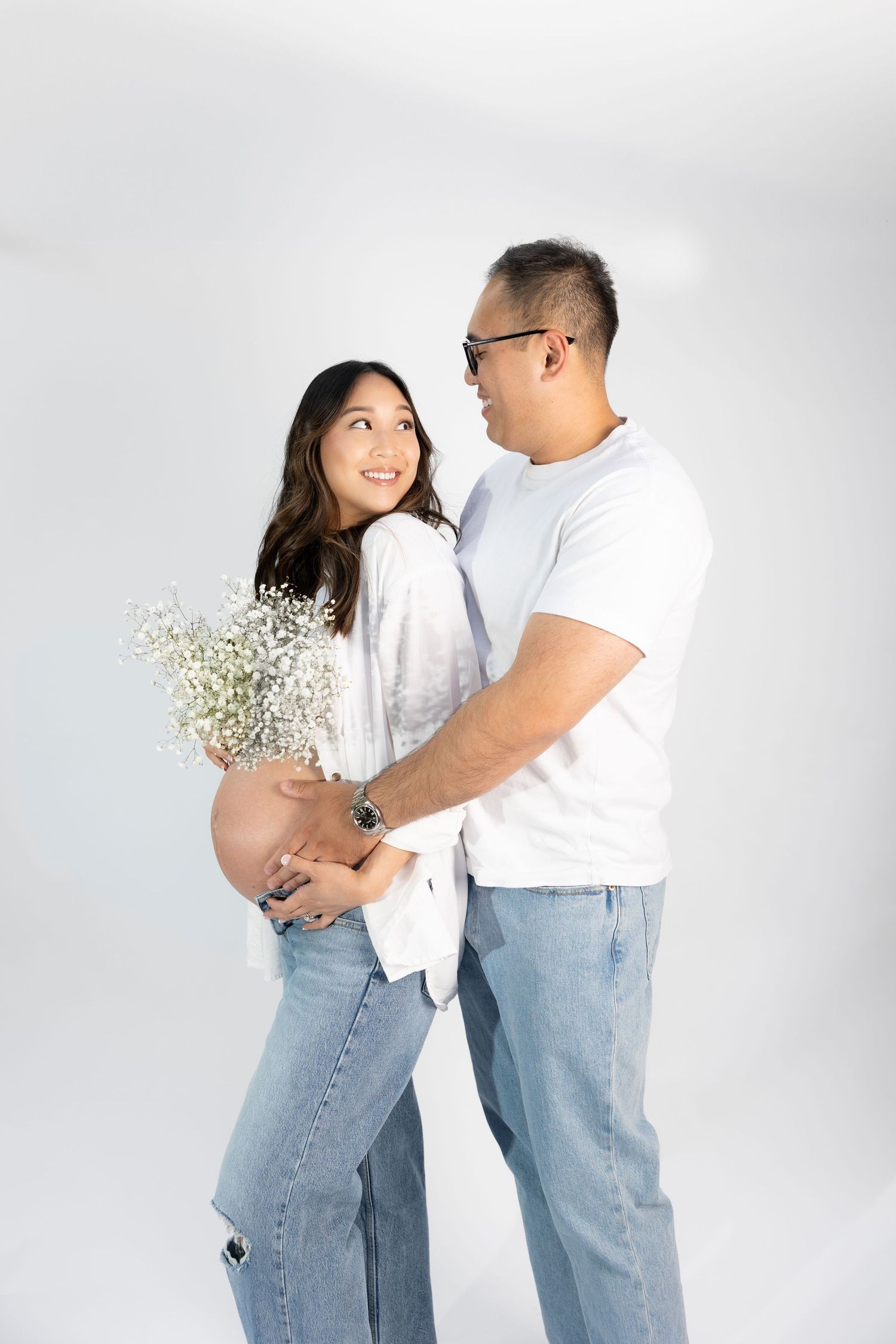 Maternity portrait of couple in white clothing against light background.