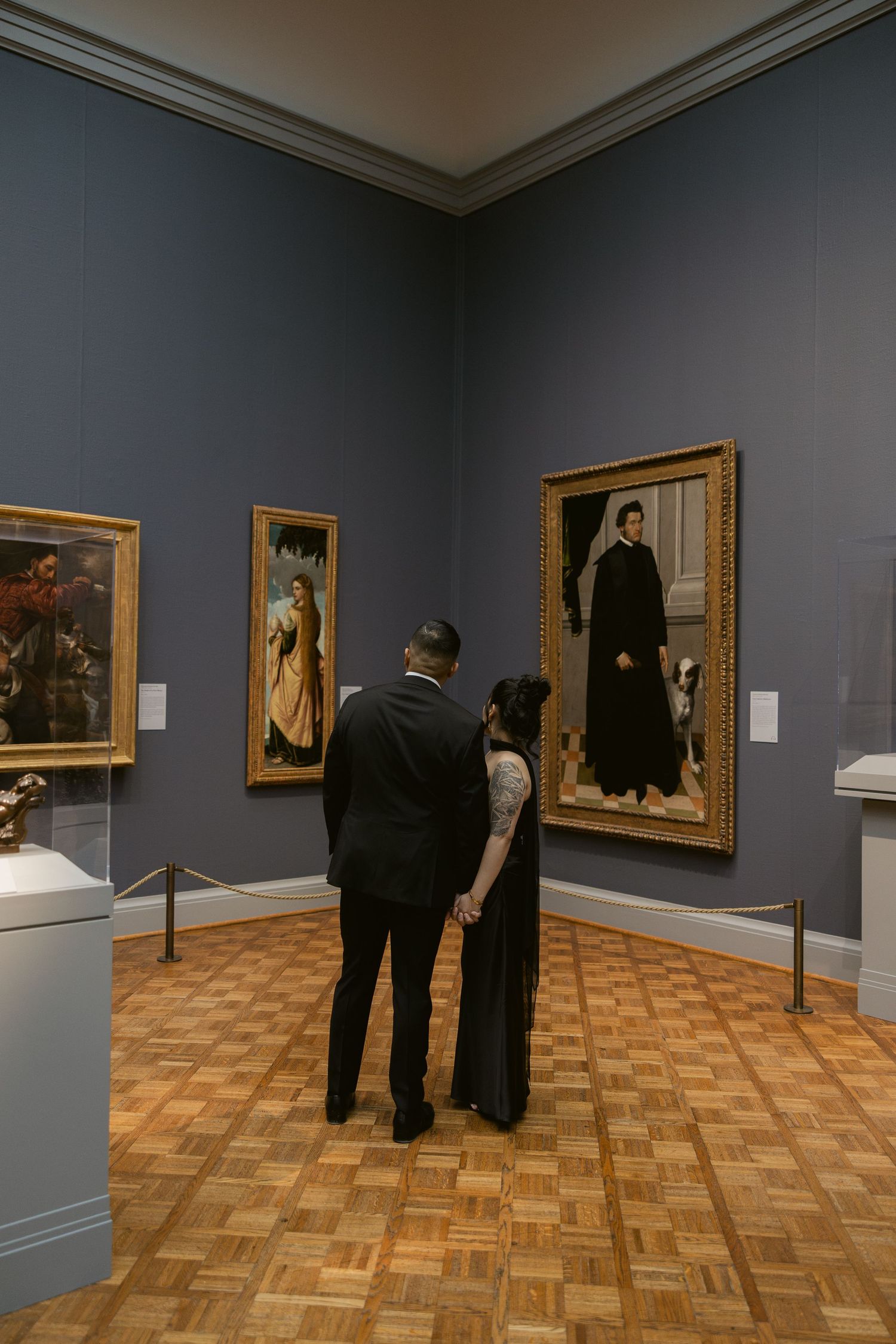 A couple admires classical paintings in a museum gallery with grey walls and wooden flooring.