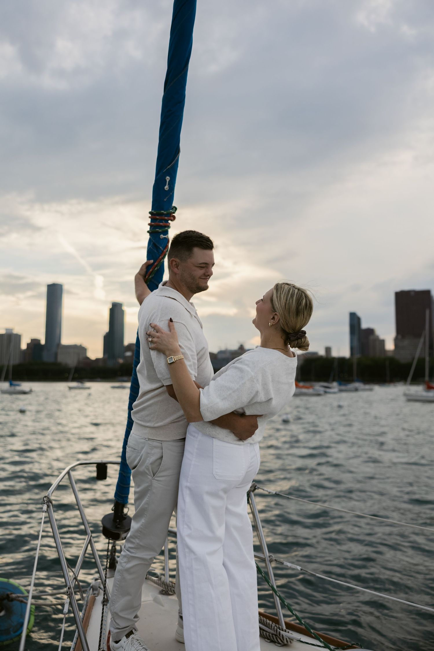 A couple in white clothing stands together on a sailboat deck with a waterfront city view.
