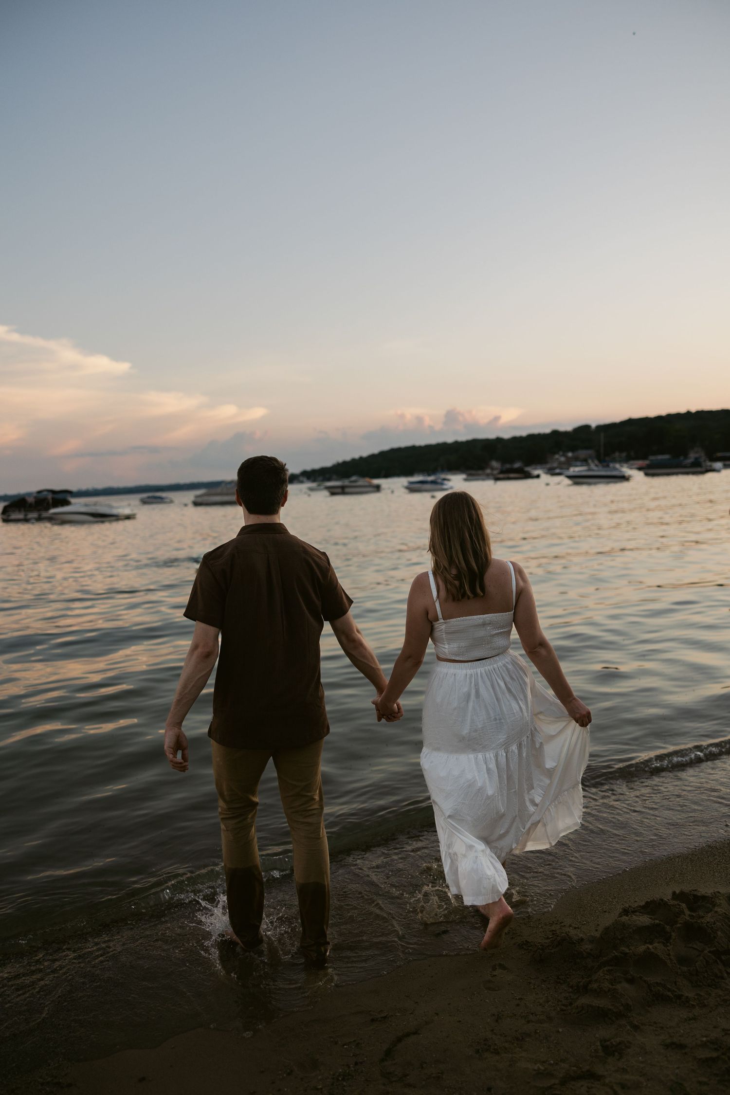 A couple wades into calm ocean waters at sunset with boats in the distance.