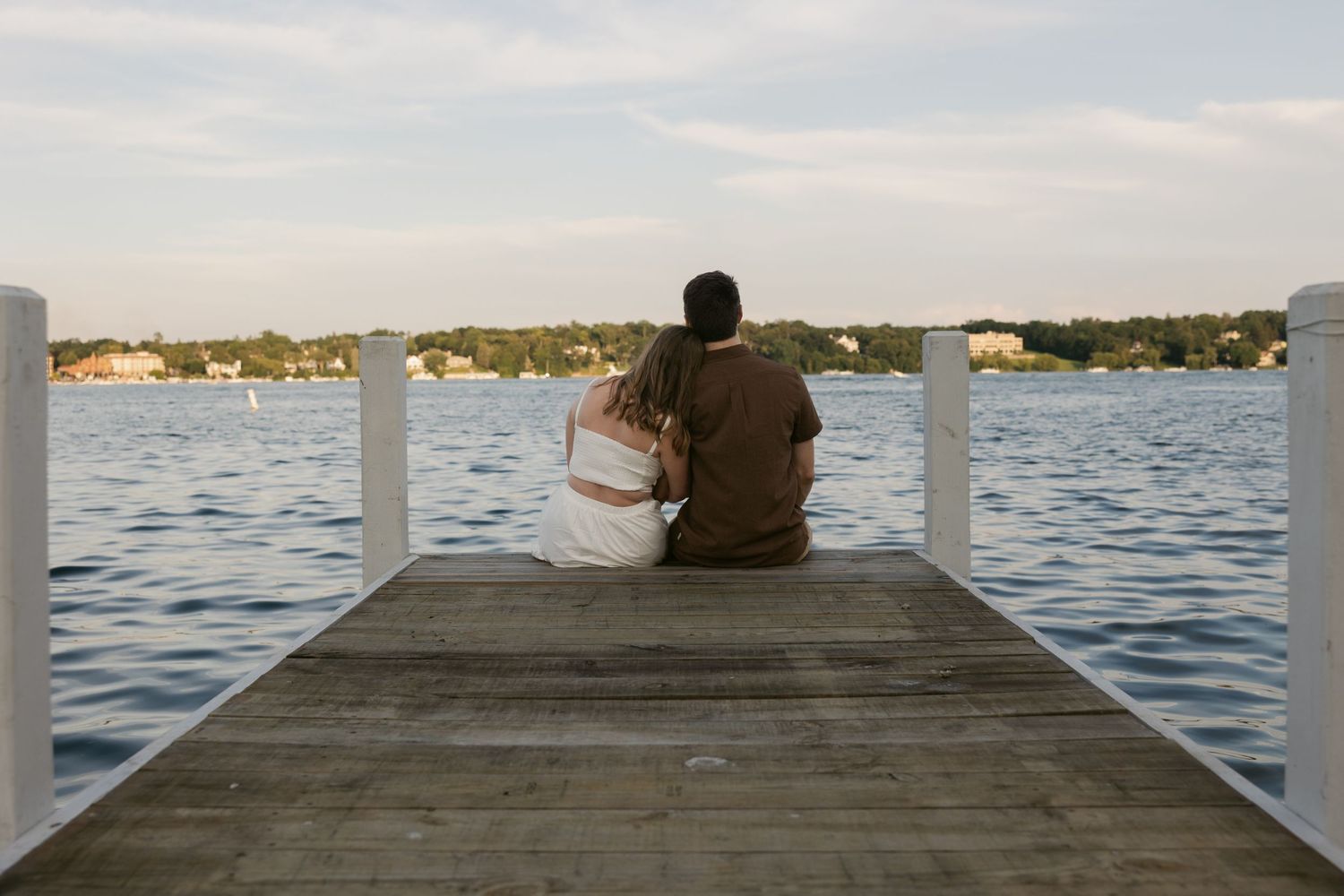 Silhouetted figure sits on wooden pier overlooking calm lake waters at sunset creating peaceful waterfront scene.