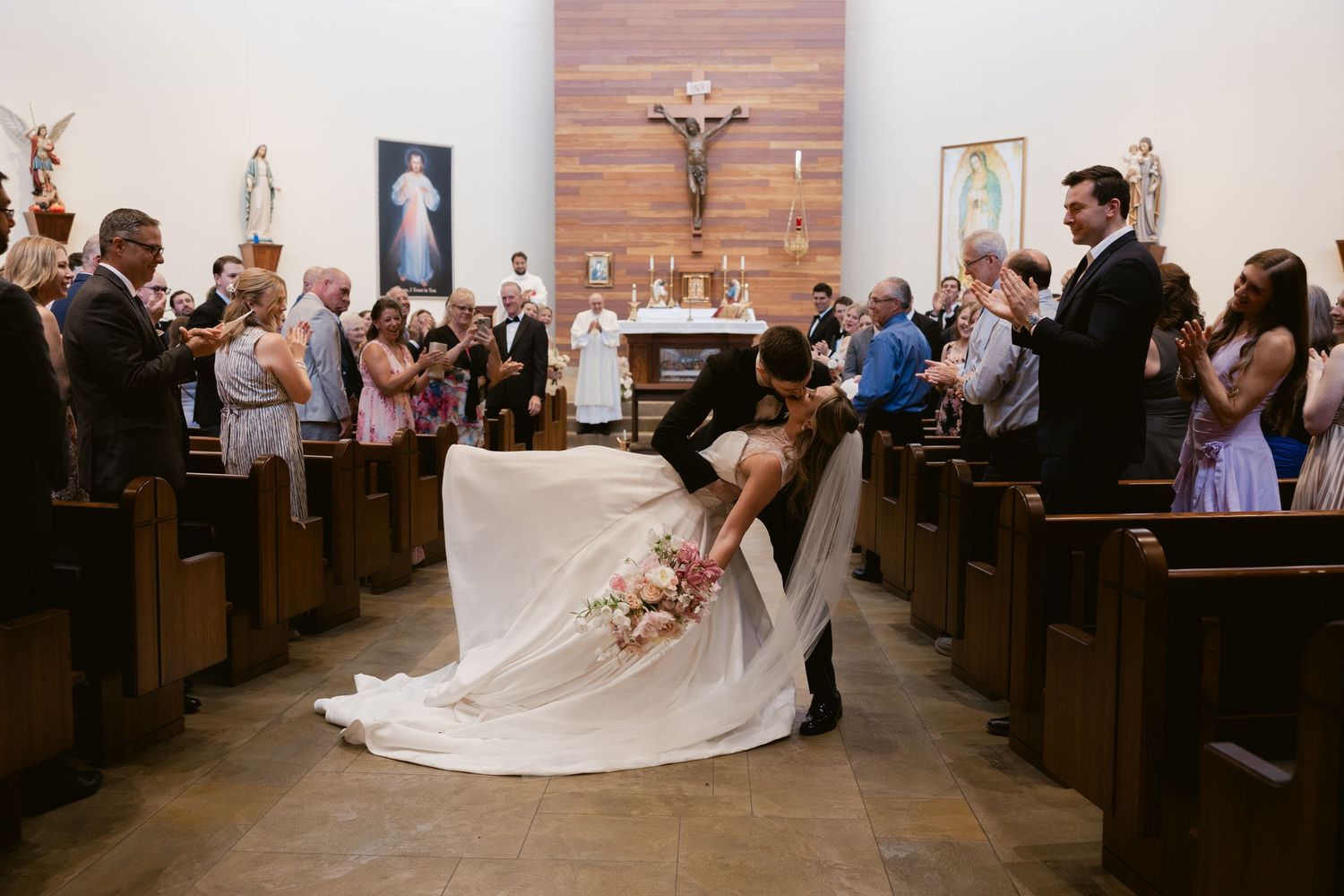 A couple performs a romantic dip pose in a church aisle surrounded by cheering wedding guests.