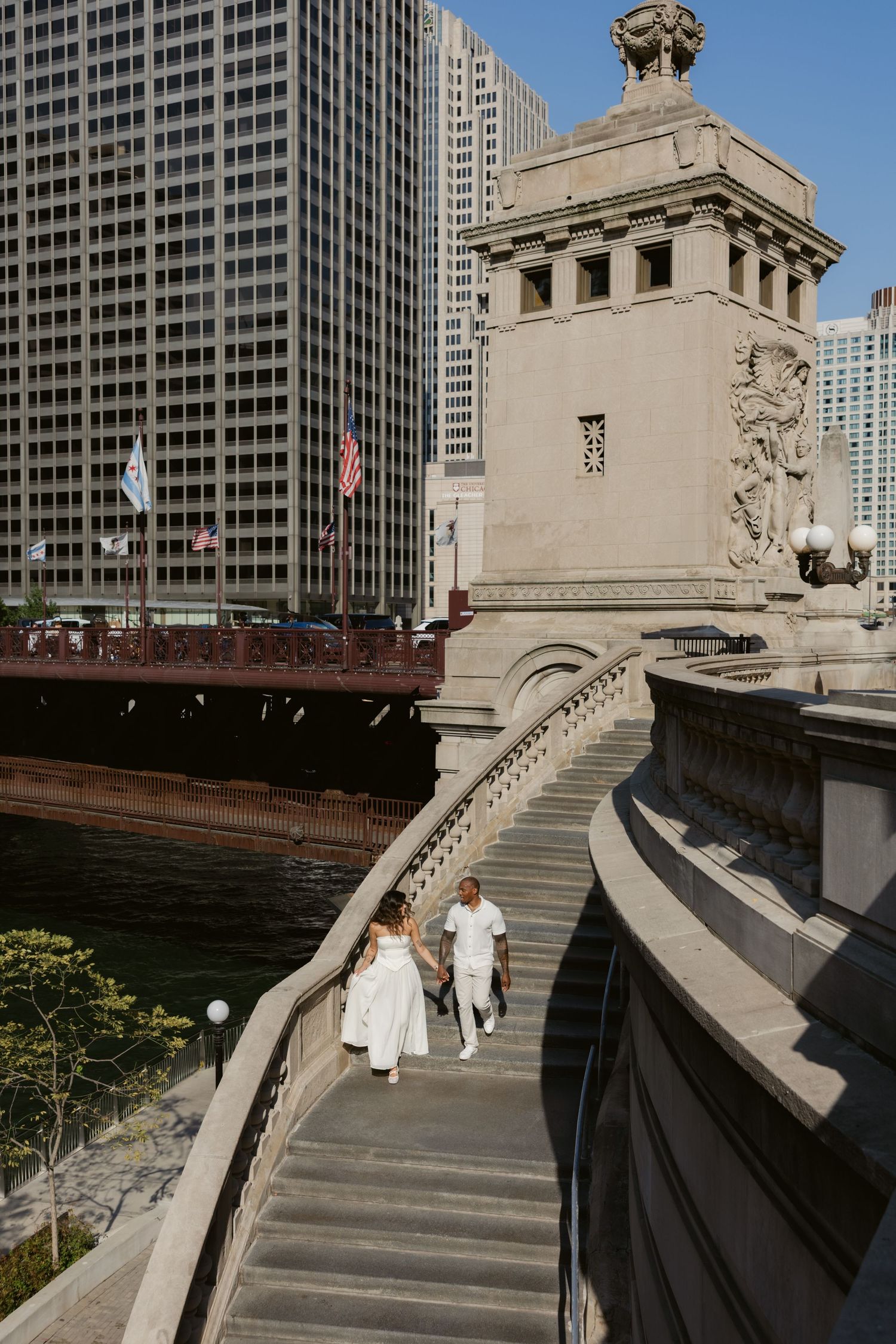 Couple in white outfits walking along ornate concrete steps with city buildings in background.