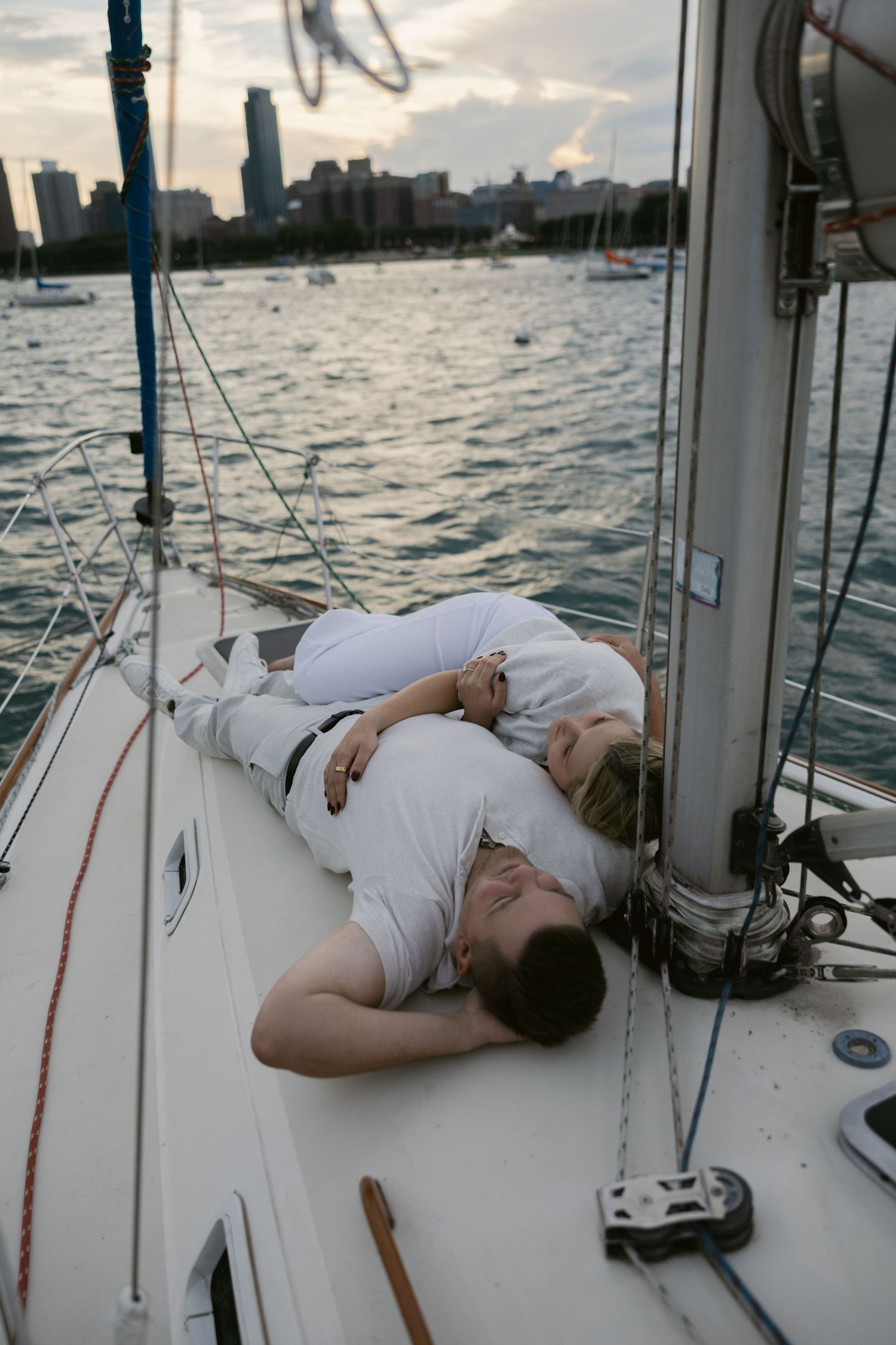 Two people in white clothing laying on the deck of a sailboat during sunset.