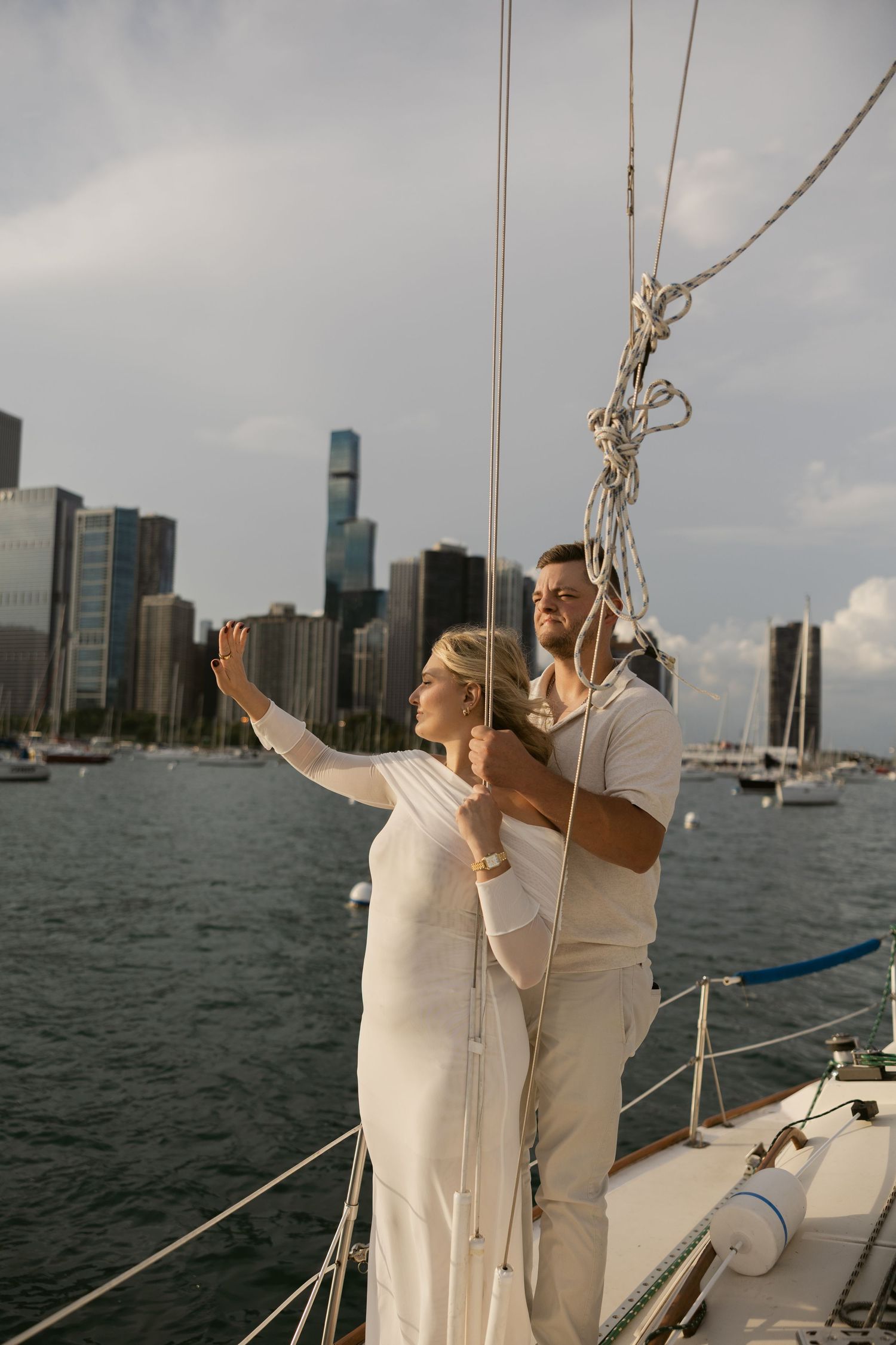 A couple in white outfits stands on a sailboat with the Detroit skyline visible across the water.