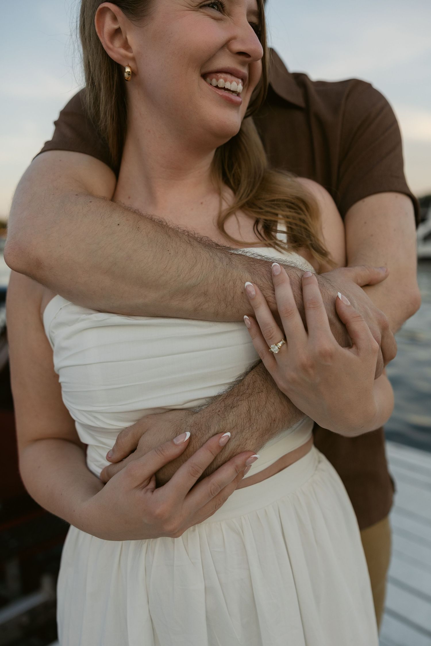 A romantic embrace on a dock shows hands clasped around a white dress, capturing an intimate moment between a couple.