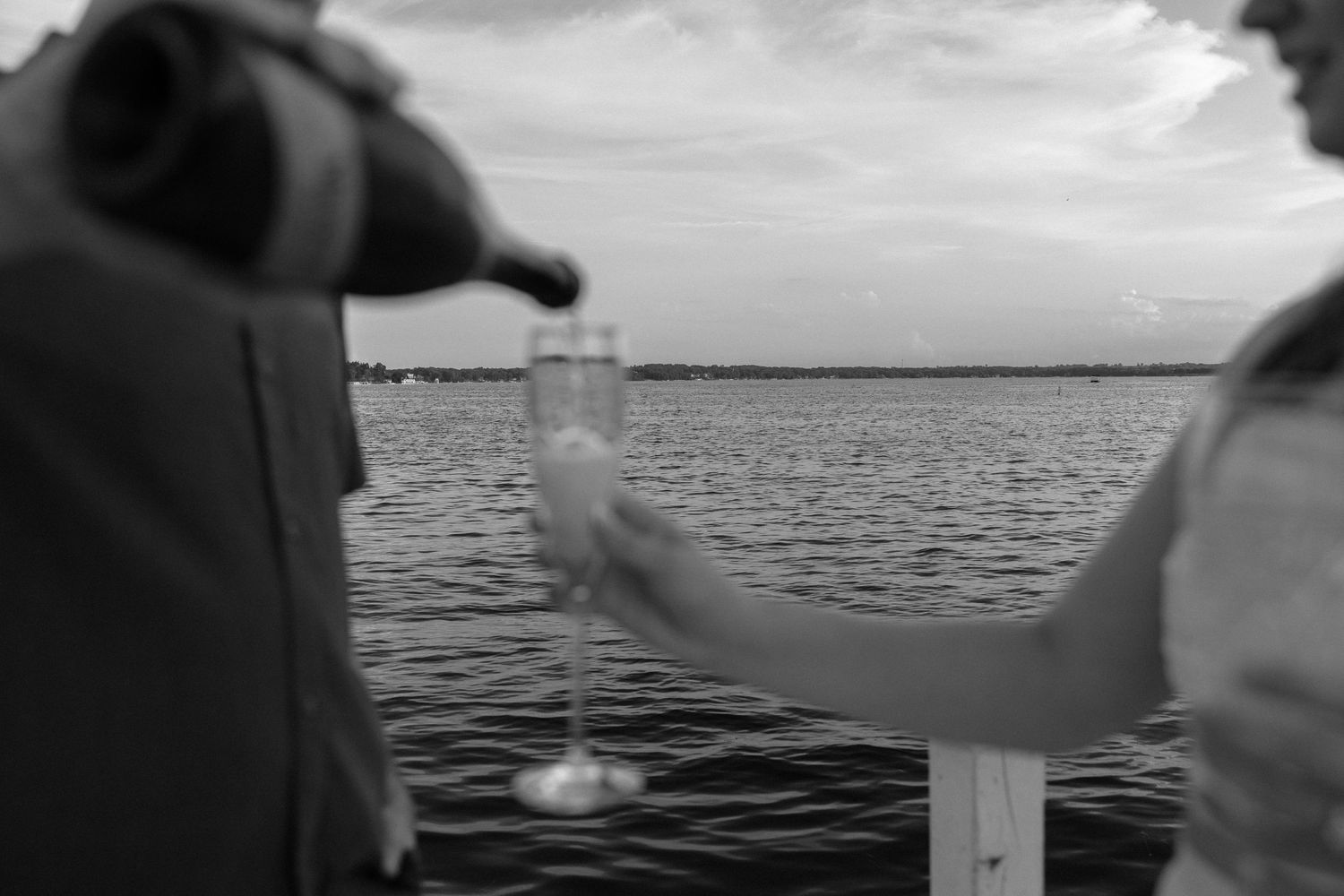 Black and white image of couple toasting with glasses on waterfront dock.