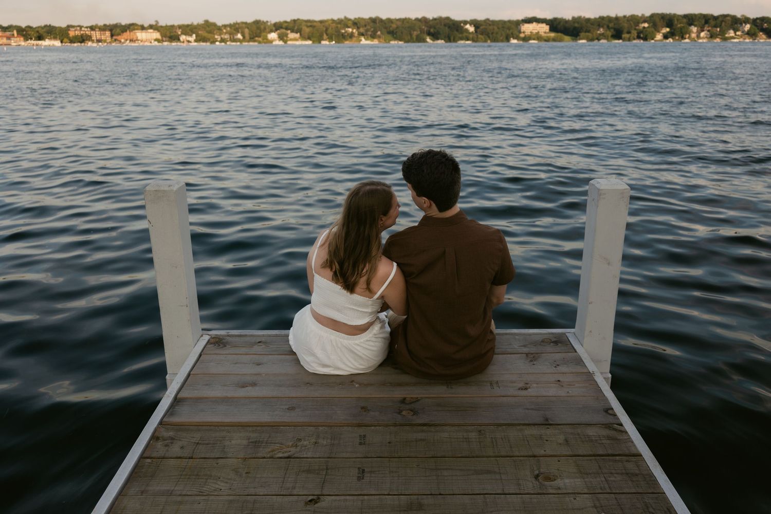 A couple sits close together on a wooden dock overlooking a calm lake at sunset.