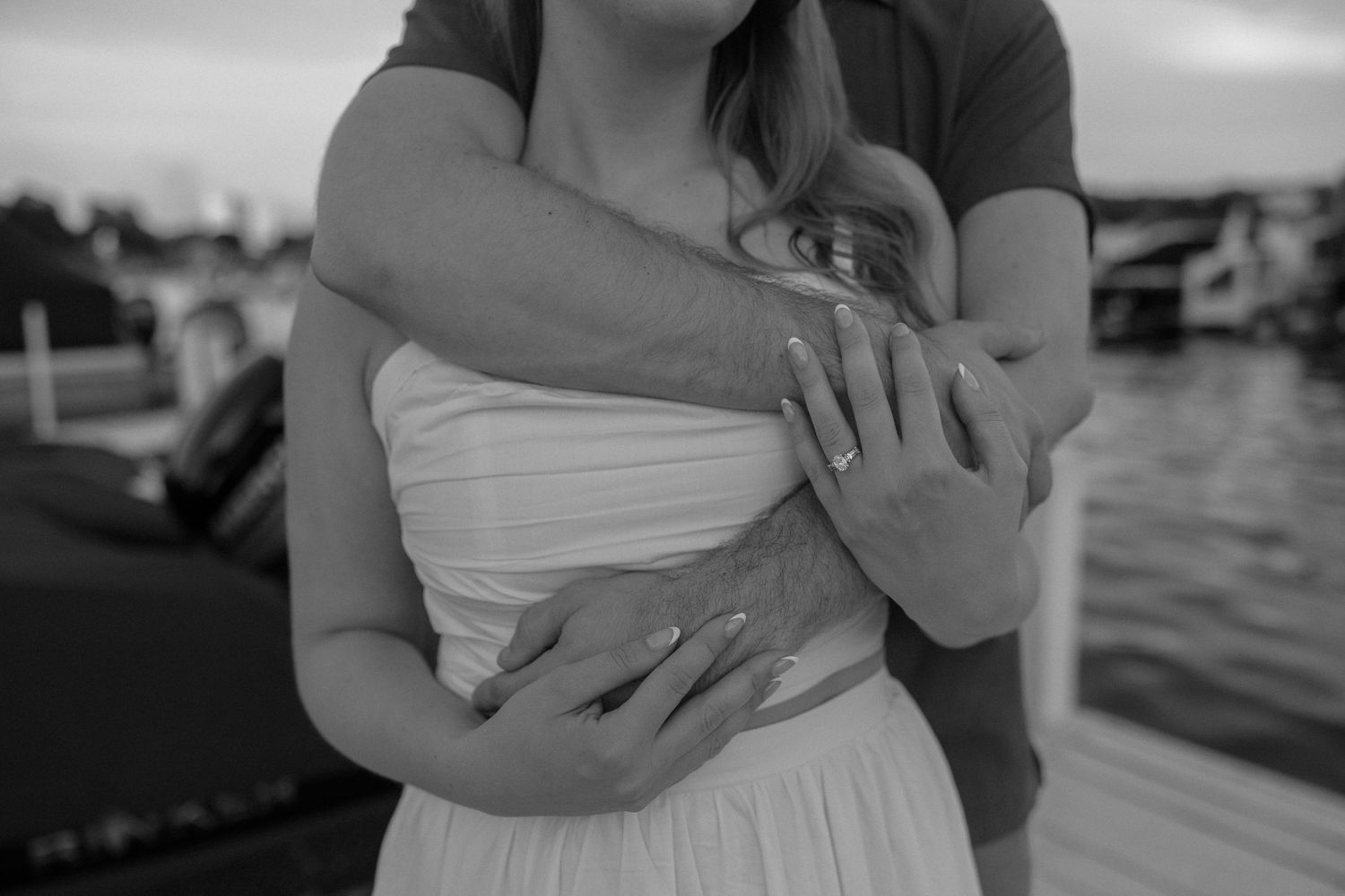 A tender black and white embrace captured near water, with a white dress silhouetted against the background.