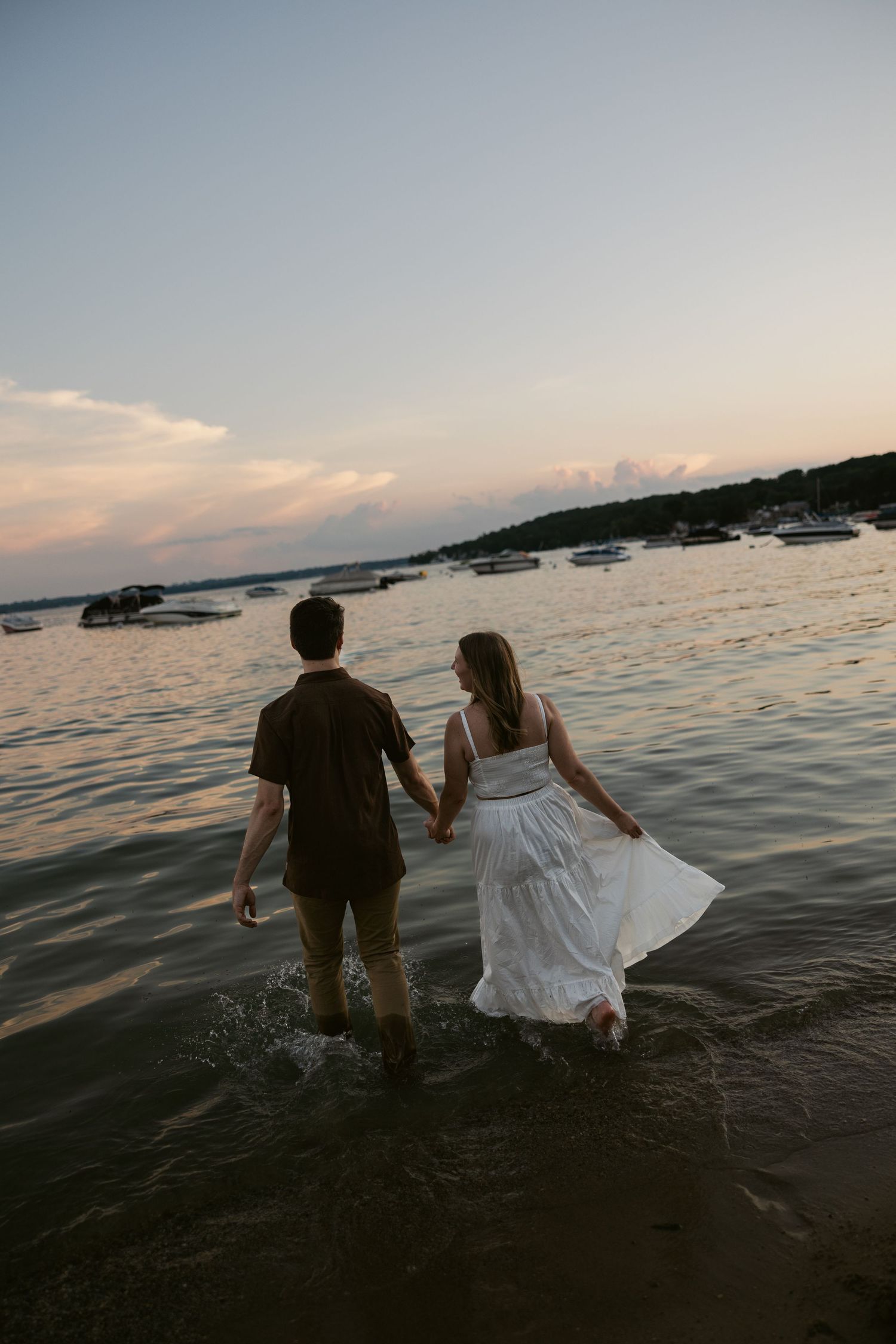 A couple splashes through knee-deep ocean water at dusk with a scenic harbor backdrop.