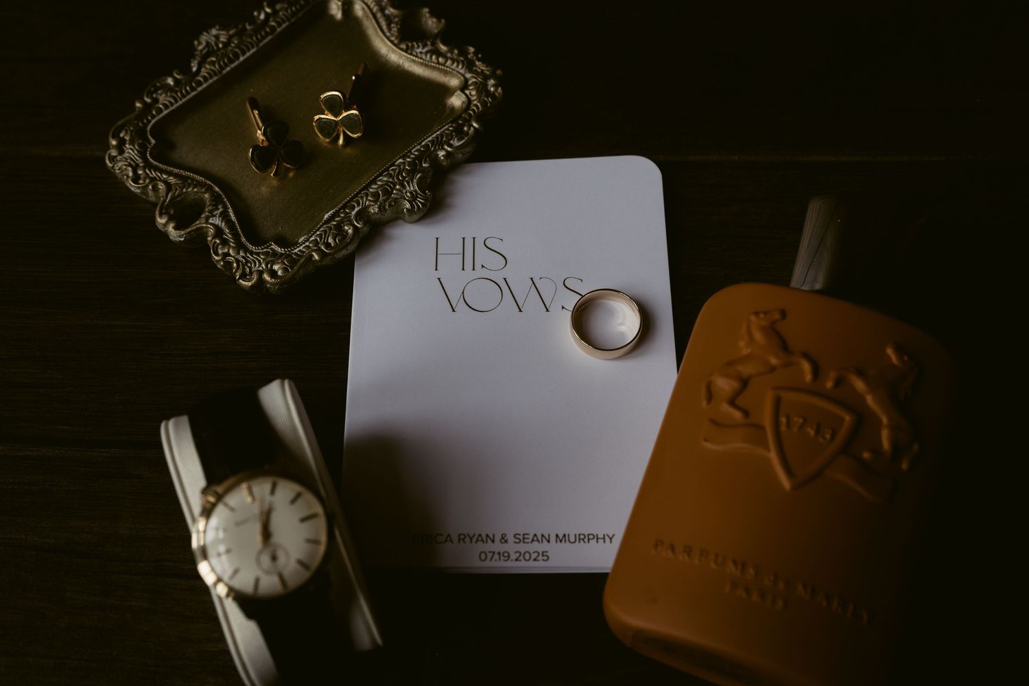 A wedding ring sits on a 'His Vows' card alongside a watch and brown leather wallet on a dark surface.