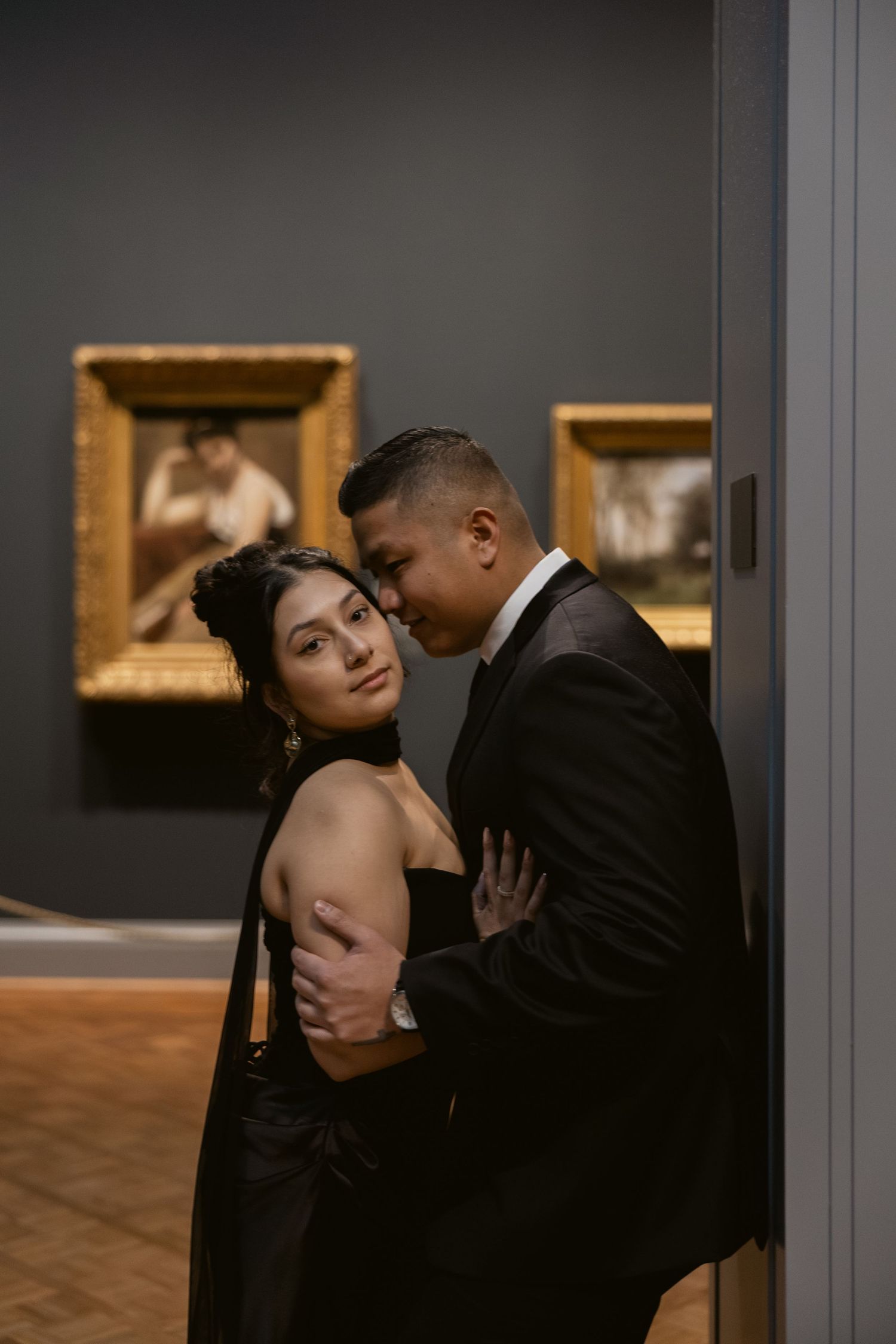 A romantic couple in formal attire share an intimate moment in an art gallery with framed paintings in the background.