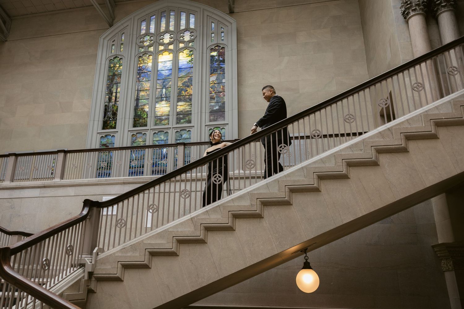A person stands on an elegant staircase with a large stained glass window illuminating the architectural space.