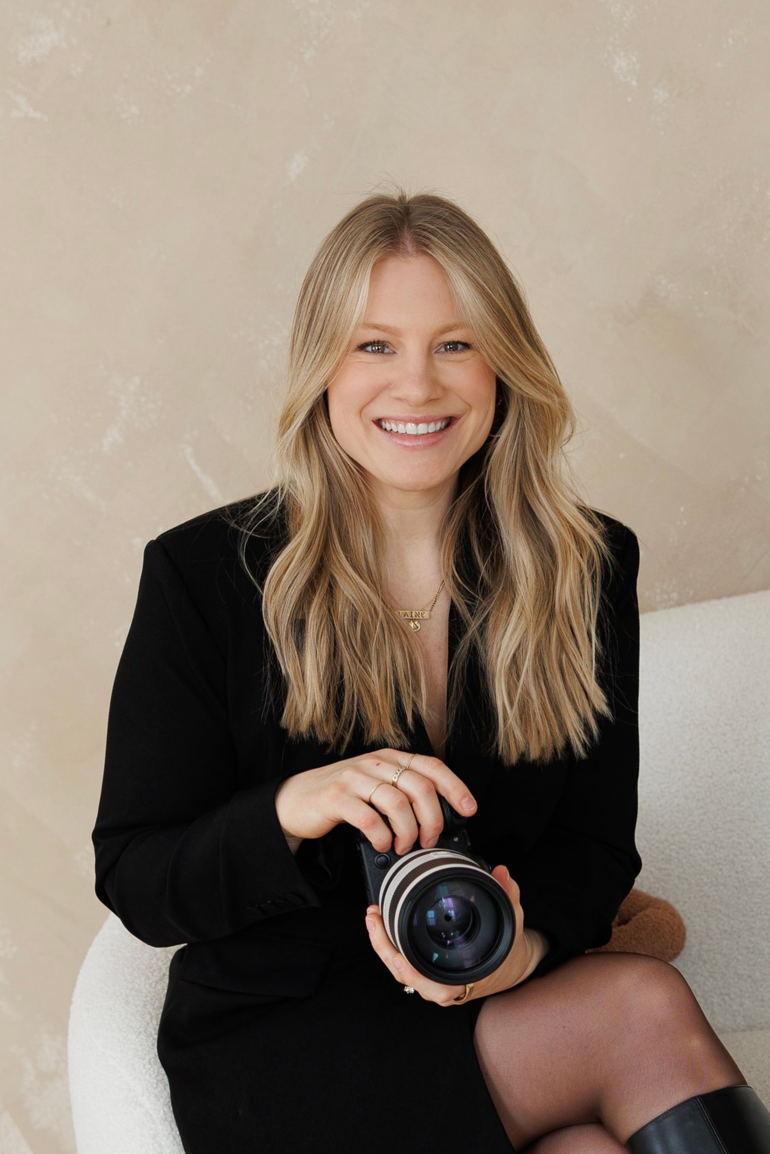 Professional photographer in black blazer poses with camera lens while sitting against beige wall in studio setting.