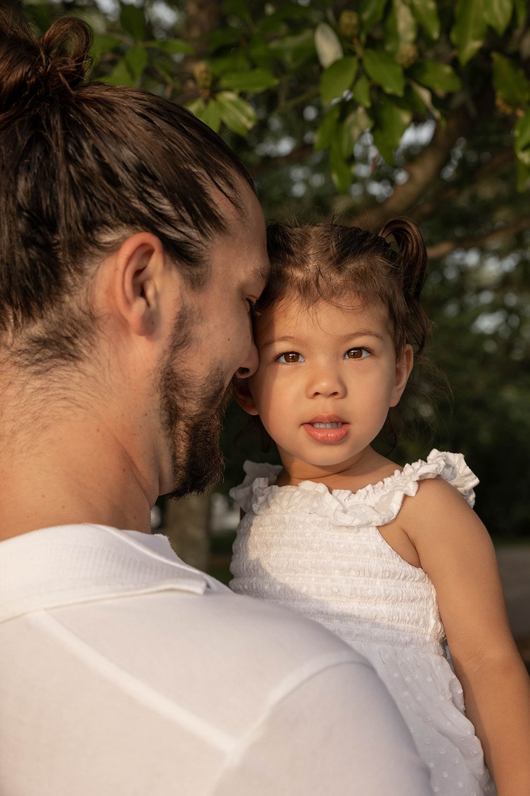 A tender moment between a caregiver in white and a young child in a white dress against a leafy background.