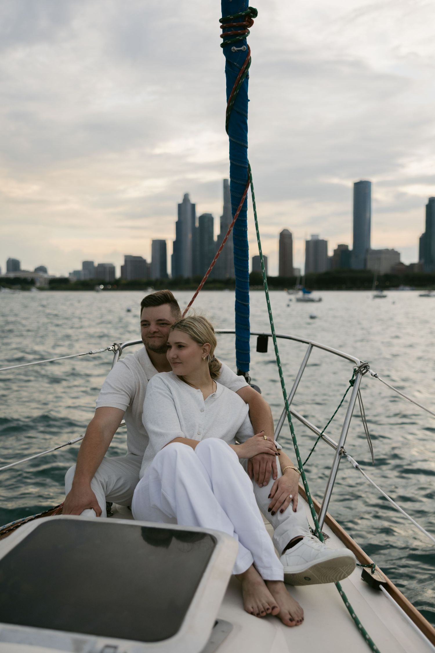 A couple relaxes on a sailboat deck with a city skyline visible in the distance.
