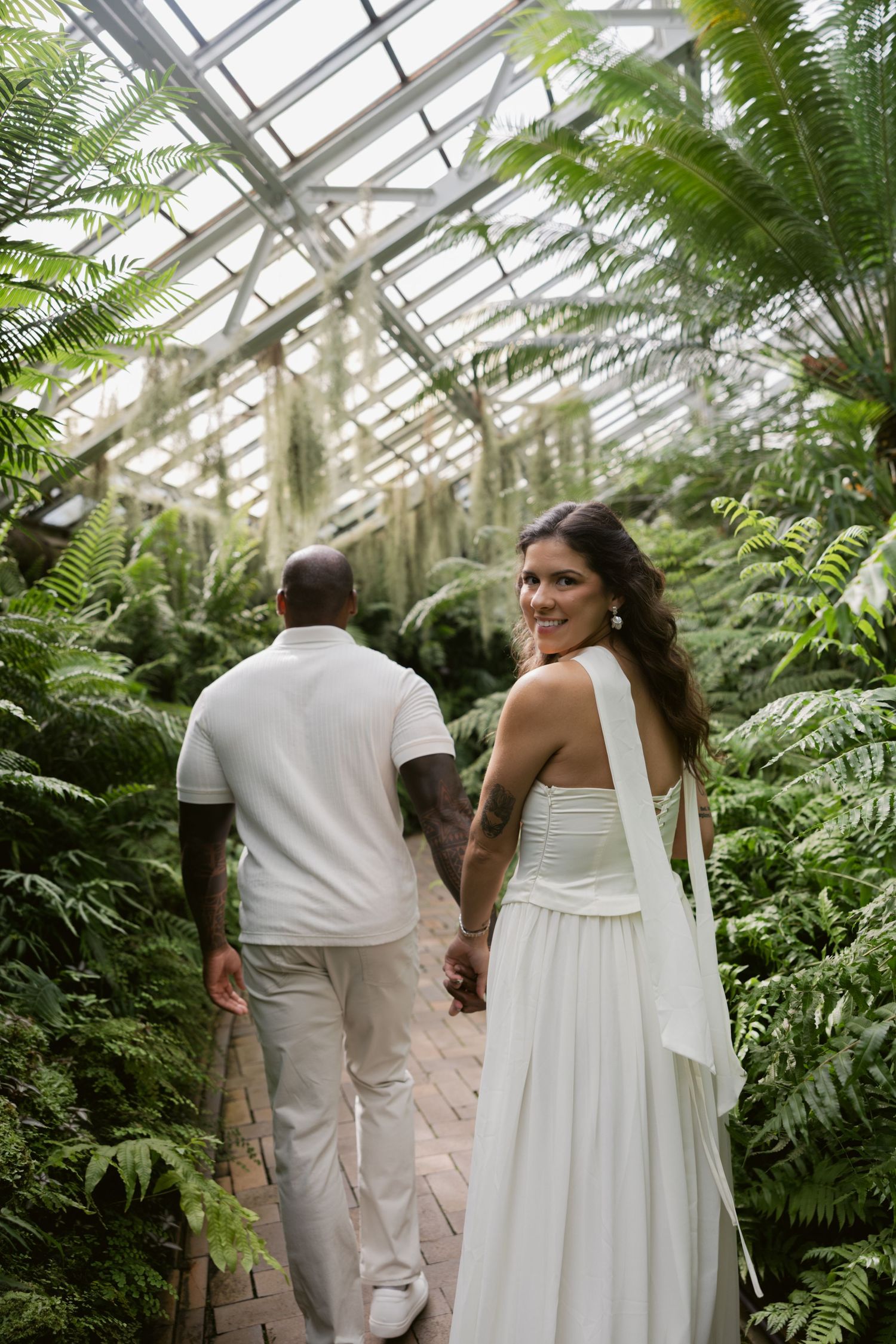 A romantic greenhouse photo session captures a couple walking among lush ferns and greenery.