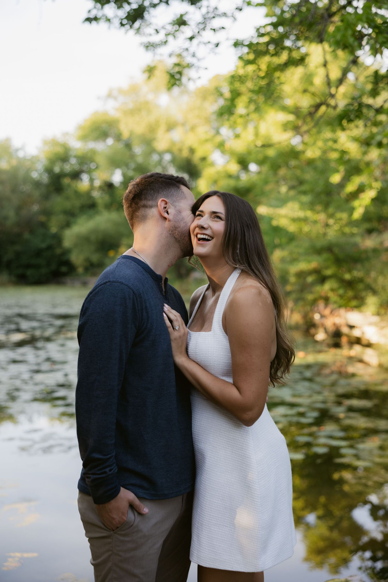 Happy couple sharing joyful moments by a pond with sunlit trees in the background.