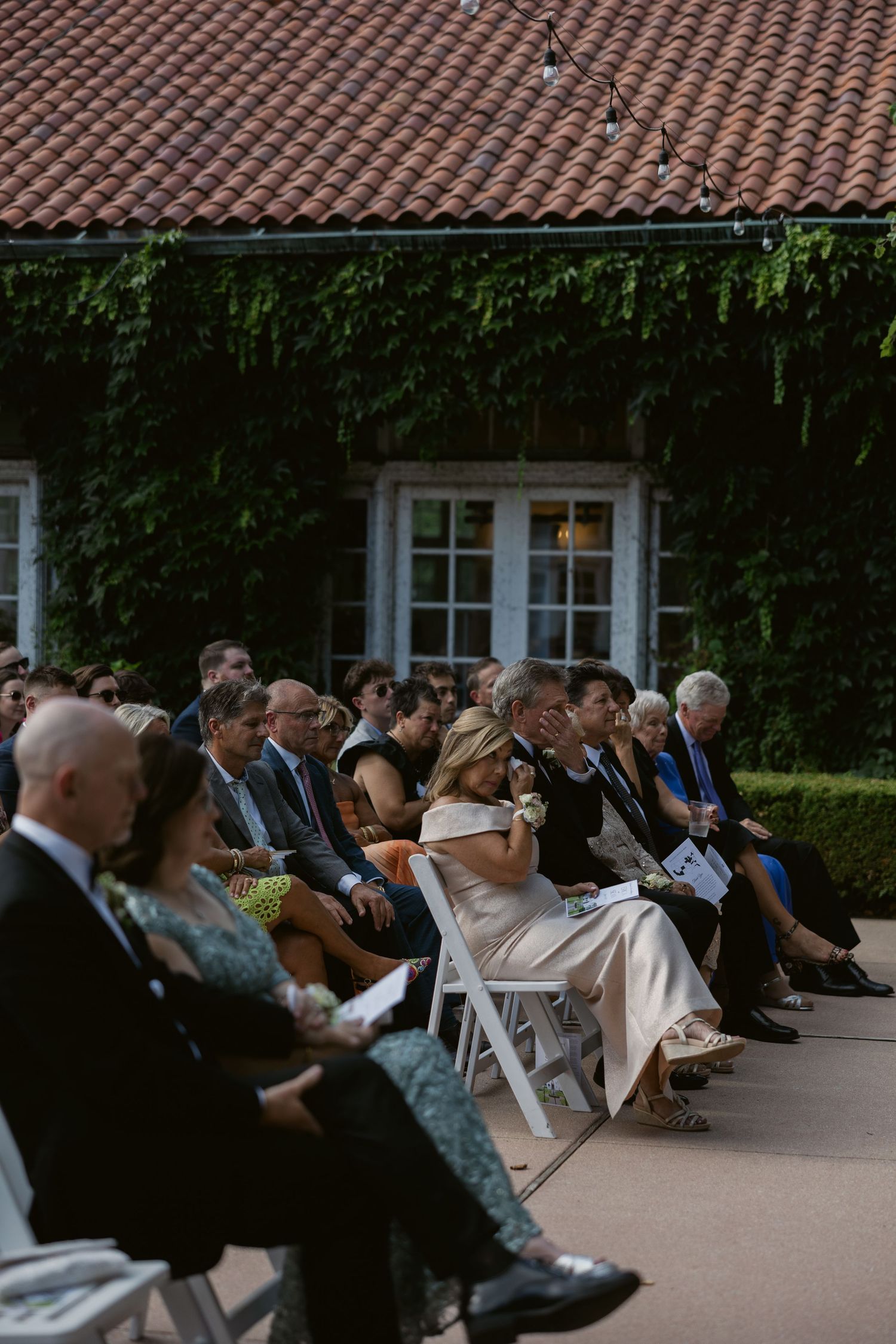 Wedding ceremony guests seated outdoors under a terracotta tile roof during a daytime service.