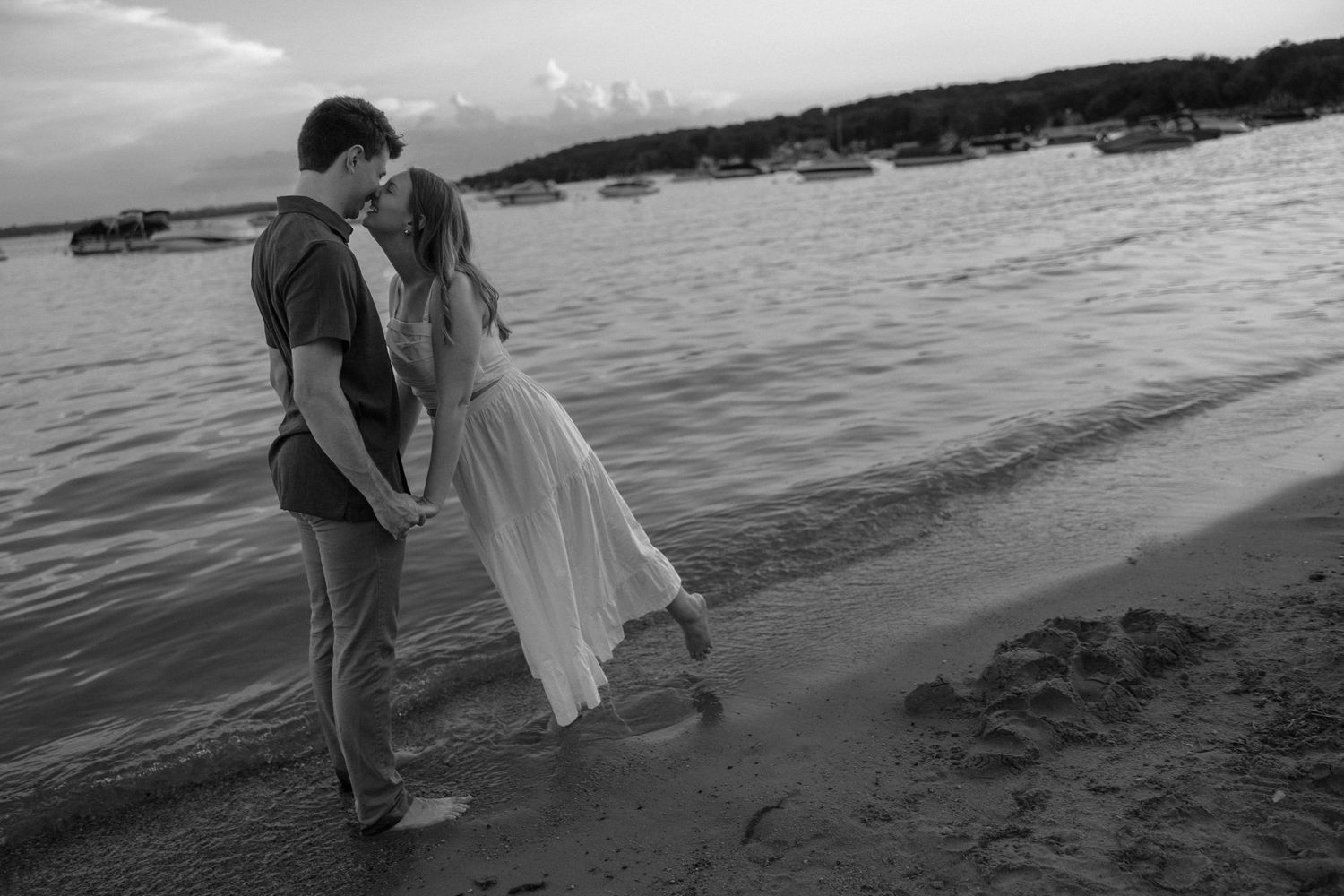 A romantic couple walks along the beach shoreline at dusk in black and white.