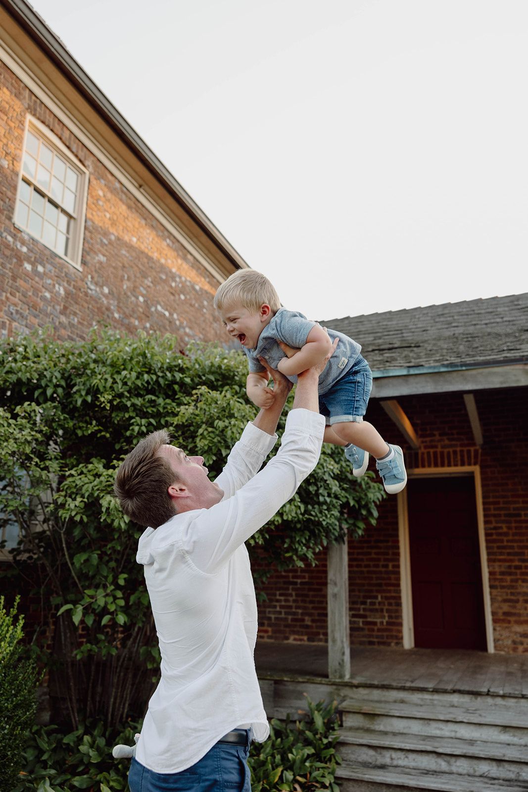A parent lifts a laughing toddler high in the air in front of a brick house.
