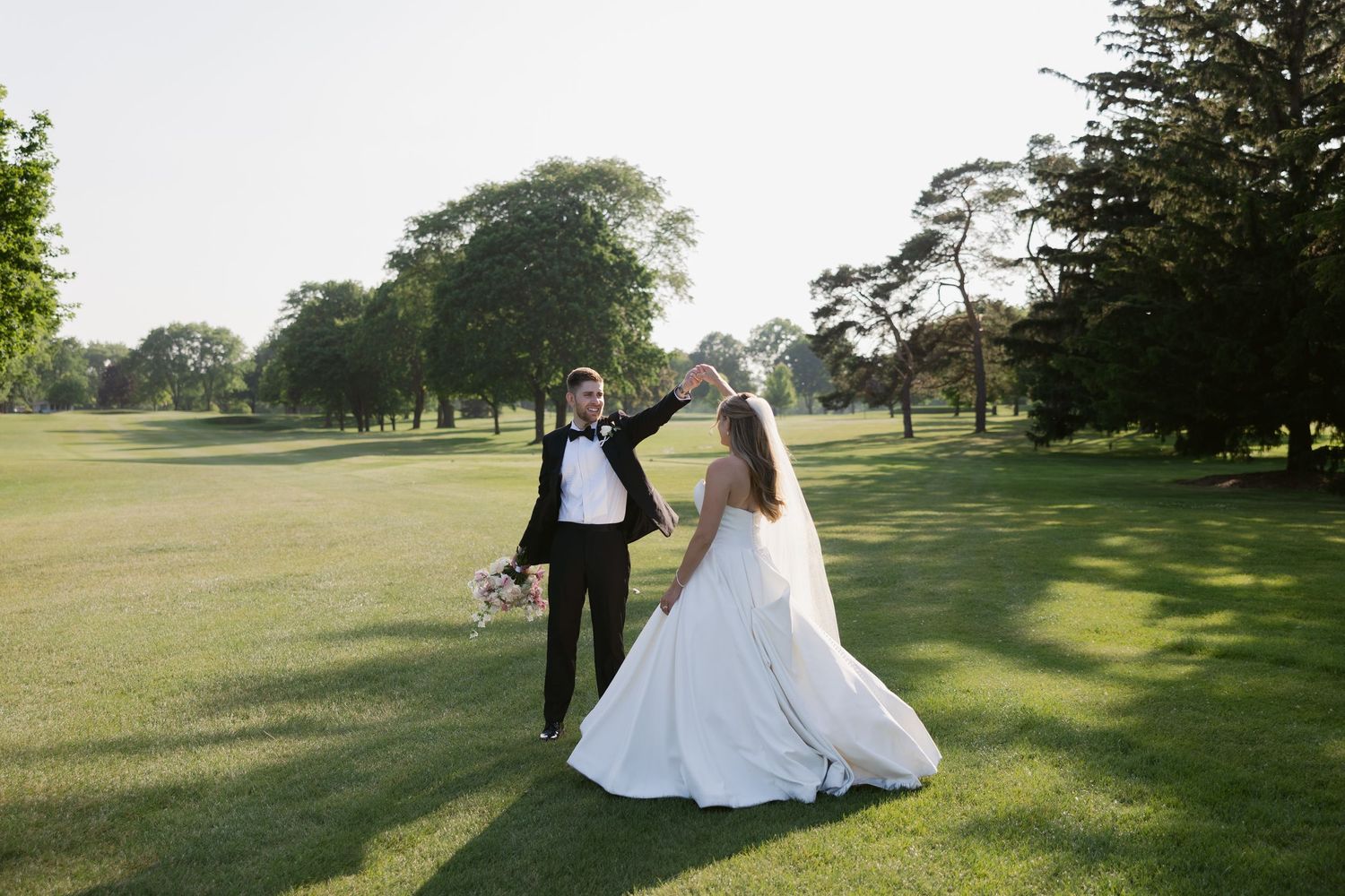 A couple in formal wedding attire walks together on a sunlit golf course during a summer celebration.