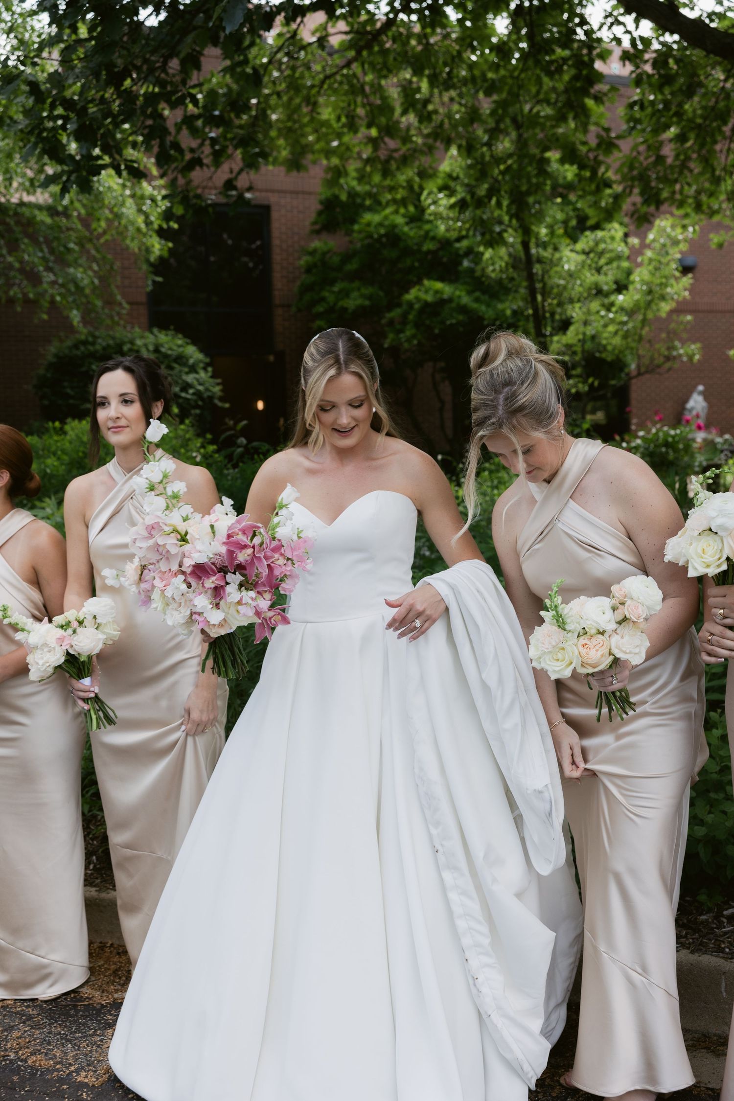 Bride in white gown poses with bridesmaids in champagne dresses holding pink and white floral bouquets.