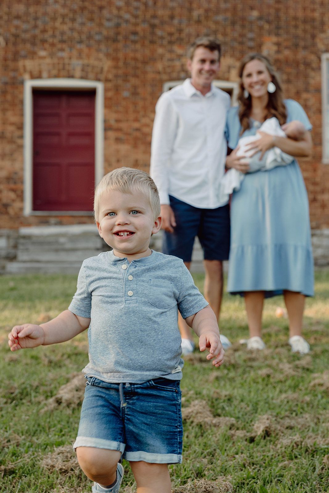 A joyful toddler runs in front of a historic brick building while parents watch from behind.