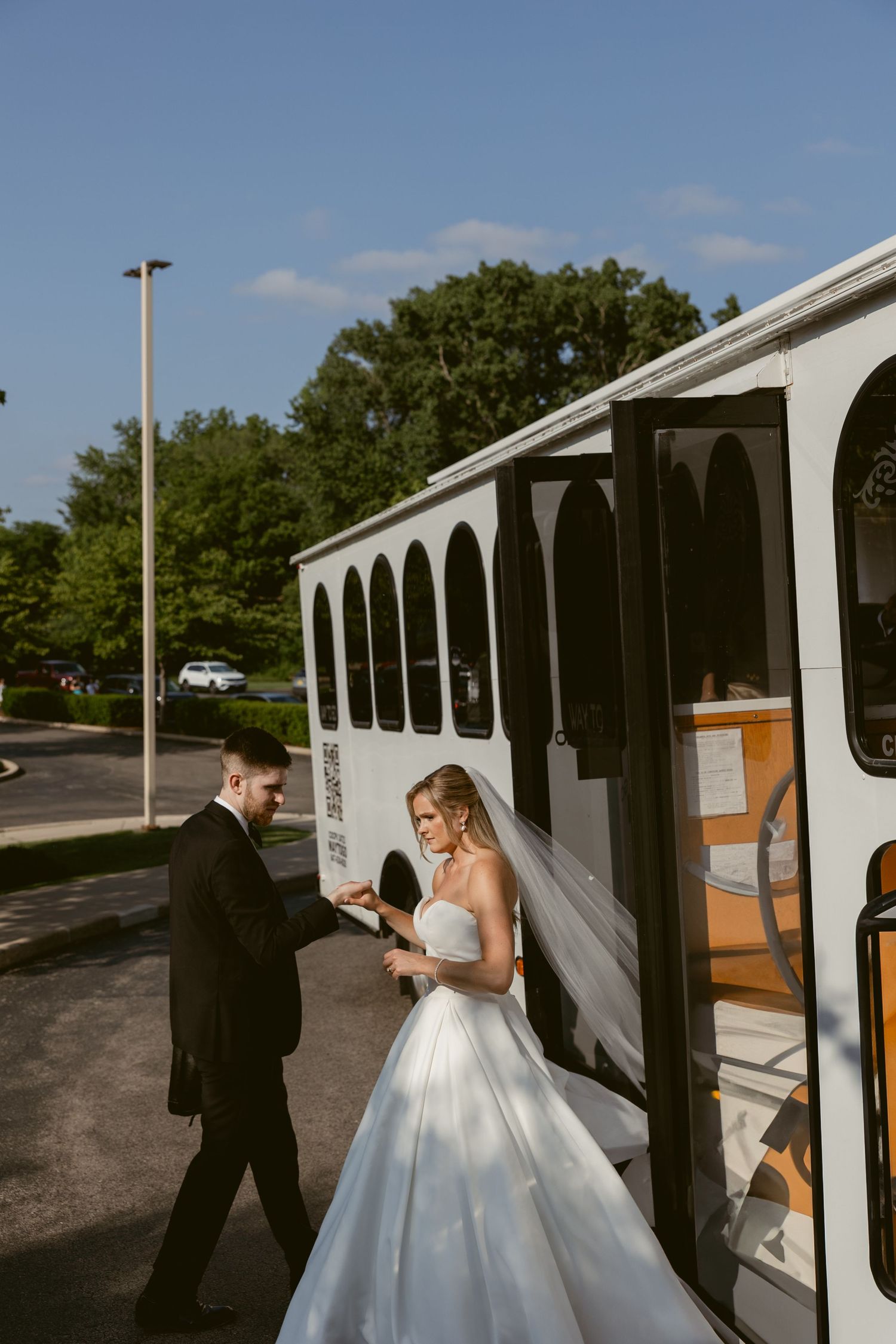 A couple boards a white shuttle bus outside on their wedding day, with the bride wearing a flowing white gown.