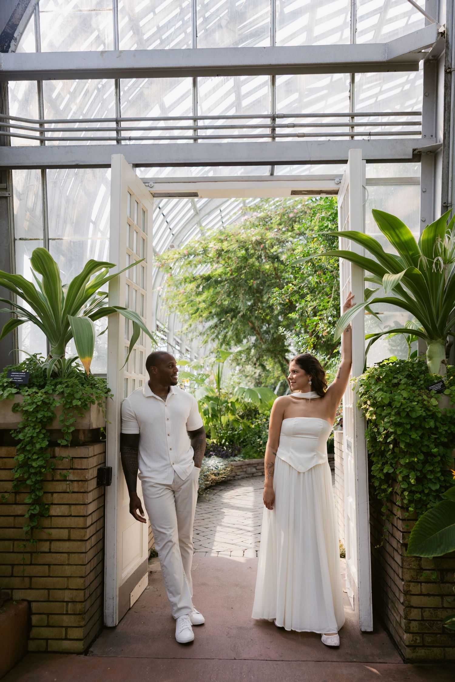 A couple in all-white outfits poses in a lush greenhouse corridor with tropical plants and brick planters.