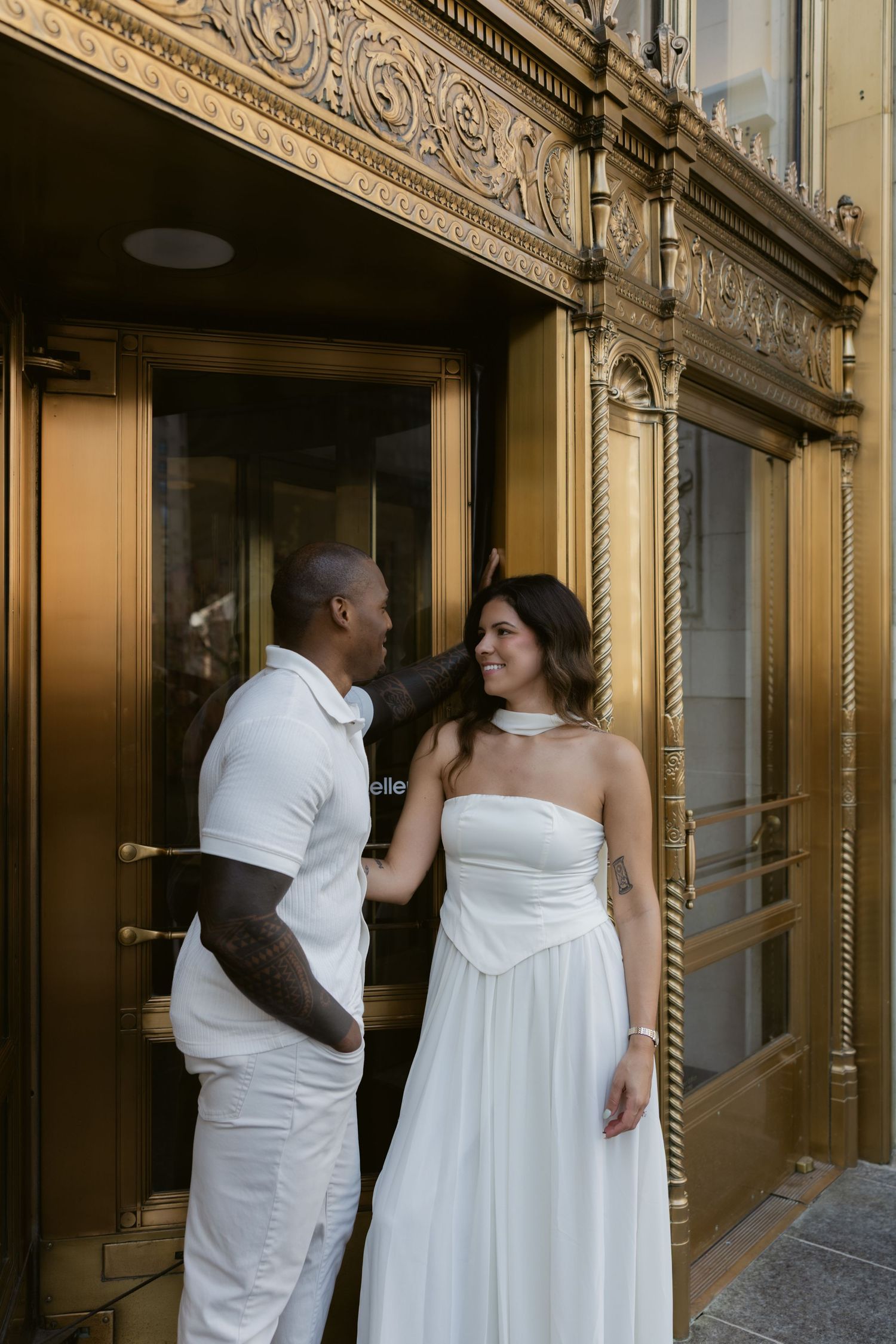 A couple in white attire poses together against an ornate vintage building facade.