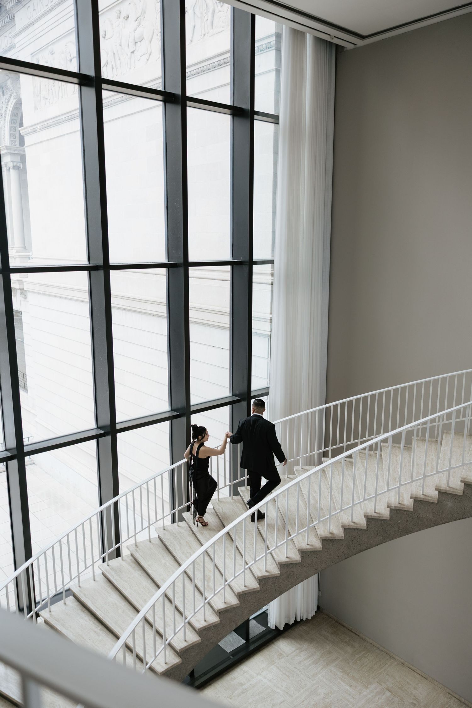 Two people in black formal wear pose dramatically on a white staircase with large windows.