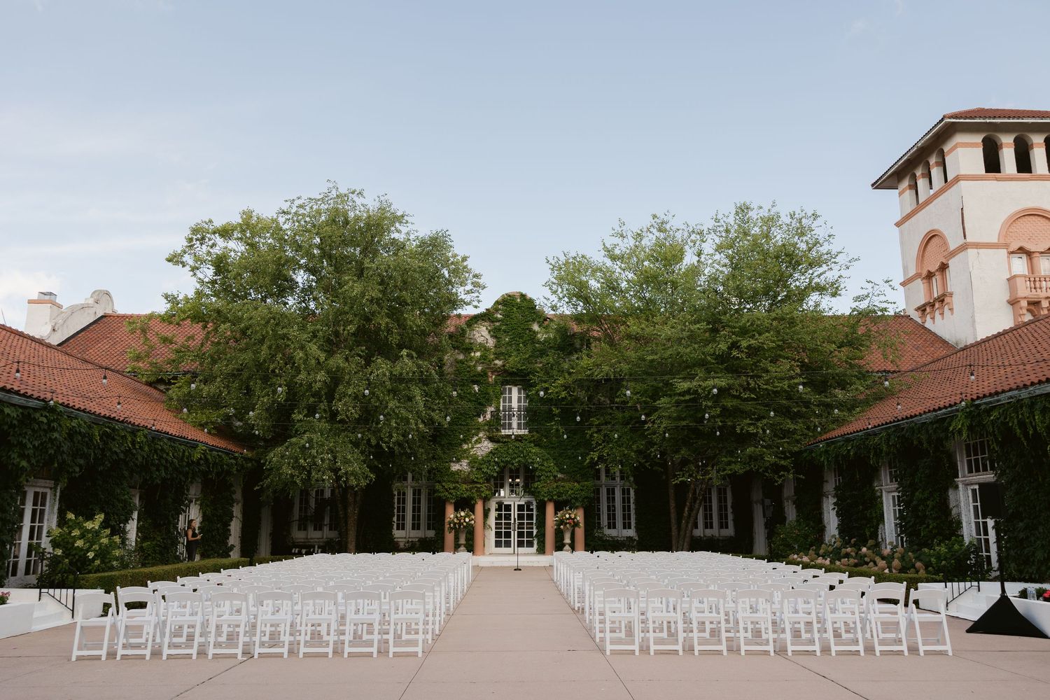 White chairs set up for an outdoor wedding ceremony in a courtyard with tall trees and Spanish-style architecture.
