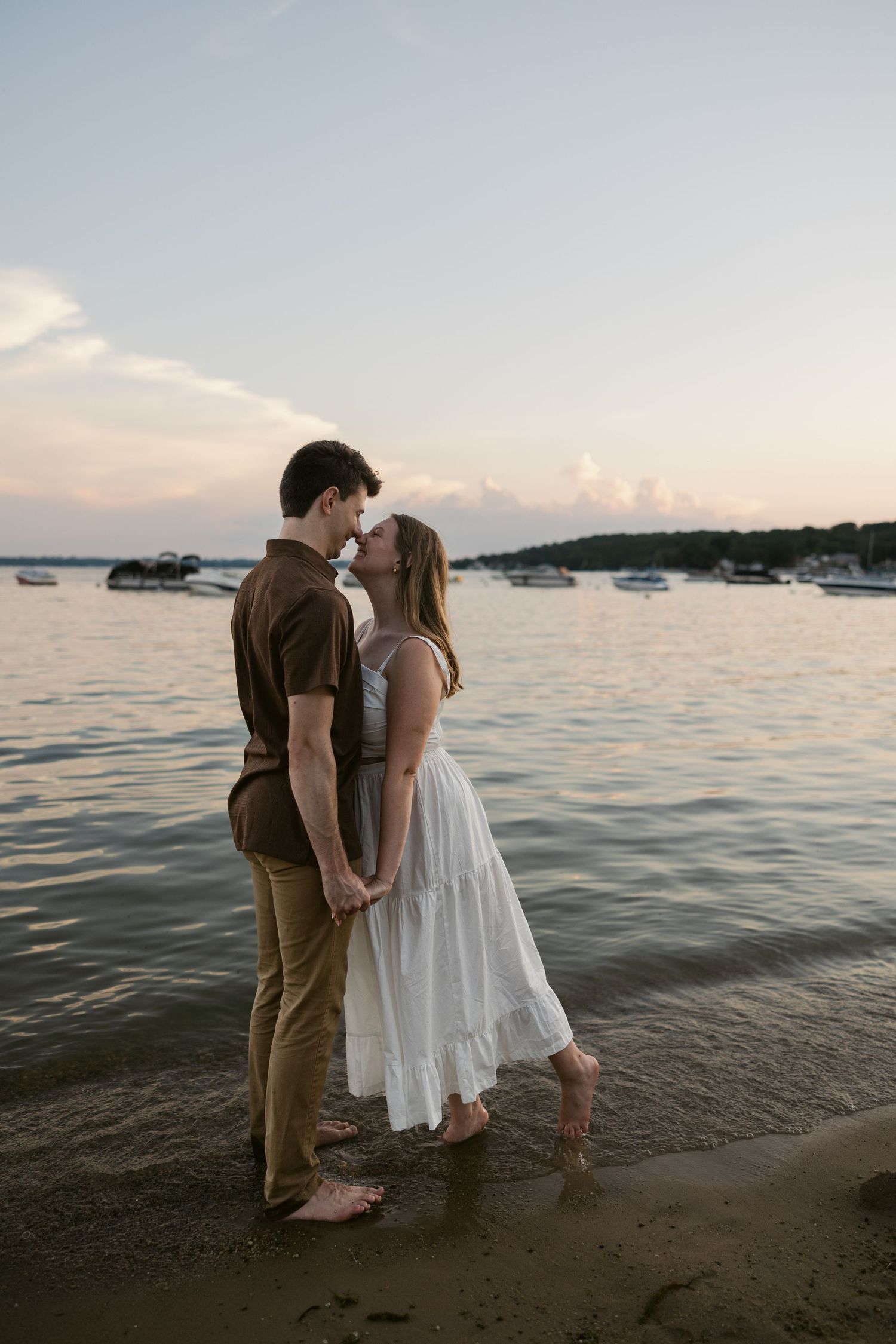 A couple shares an intimate moment on the beach at sunset with boats in the background.